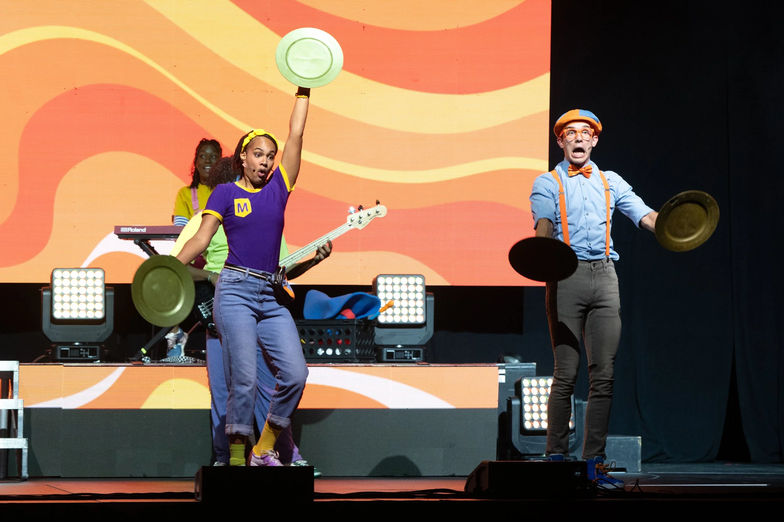 Theatrical performance with three actors on stage, two men and one woman, dressed in colorful costumes, with drumsticks, a bass guitar, and a keyboard, engaged in a lively act.