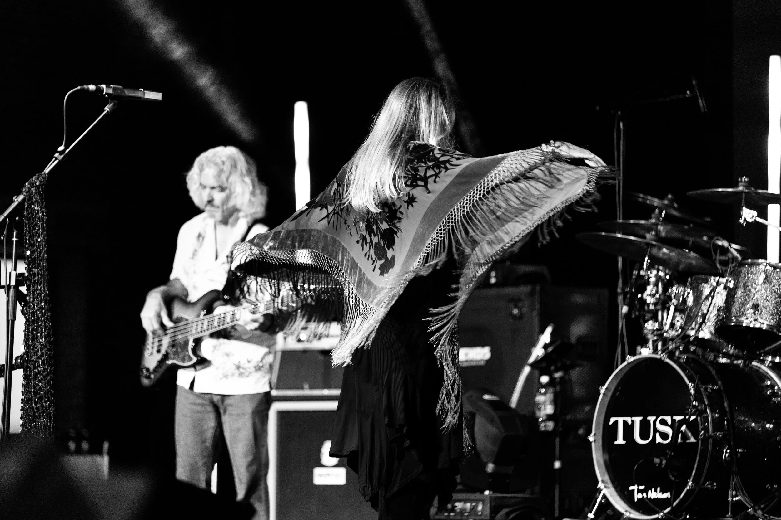 Two musicians performing on stage; one playing drums and the other with a guitar, with a featuring drum set with the word "TUSK" on the bass drum.