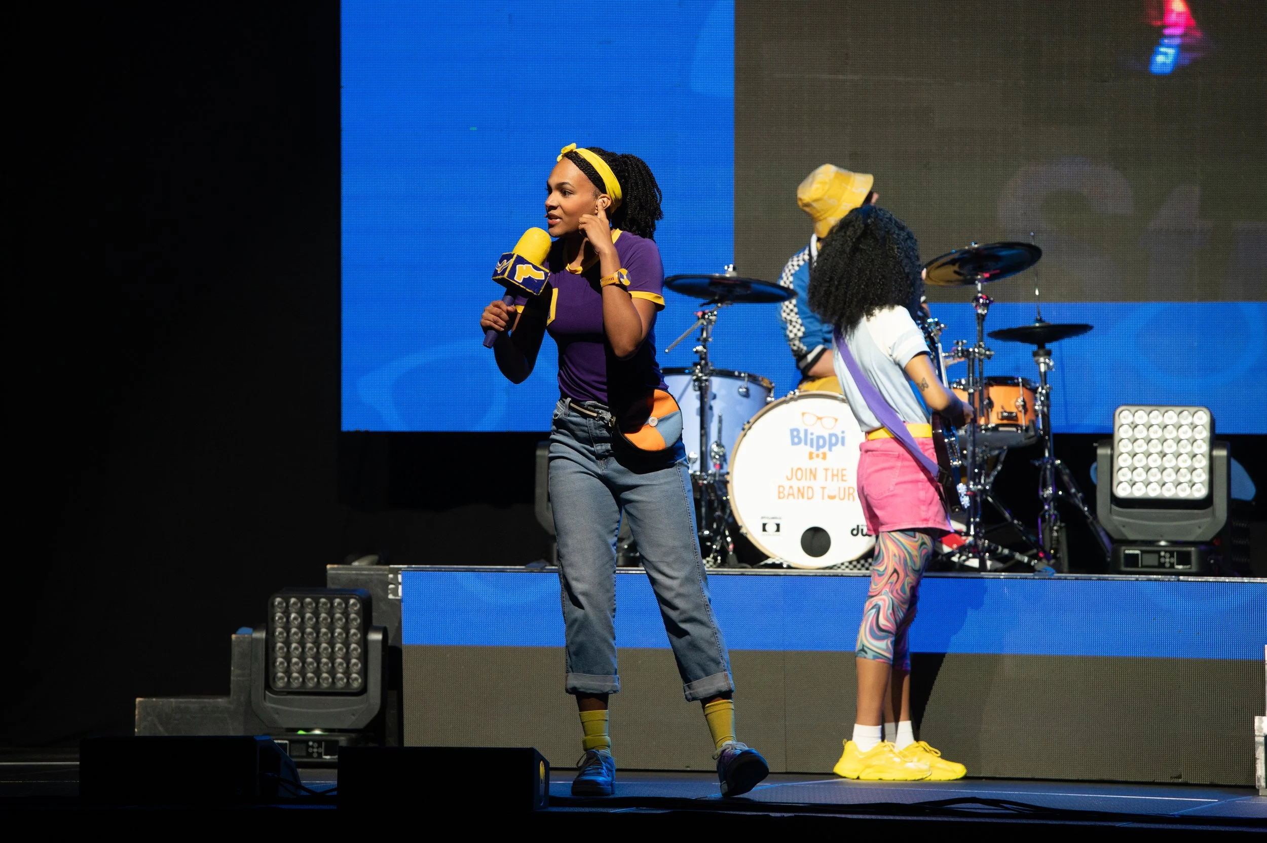 A woman with curly hair, wearing a purple and yellow outfit, holding a microphone, standing on stage with children, in front of a drum set with Blippi branding, colorful stage lights.