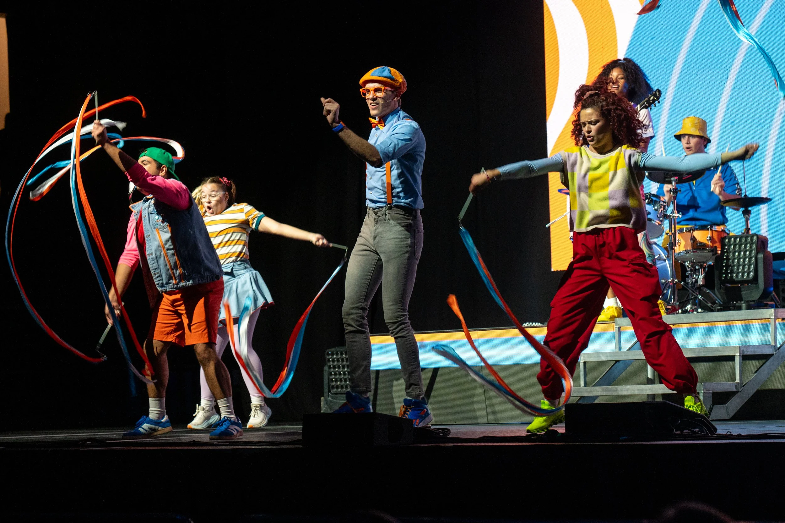Children and an adult perform on stage with ribbons, colorful clothing, and musical instruments in the background, engaging in a lively dance or performance.