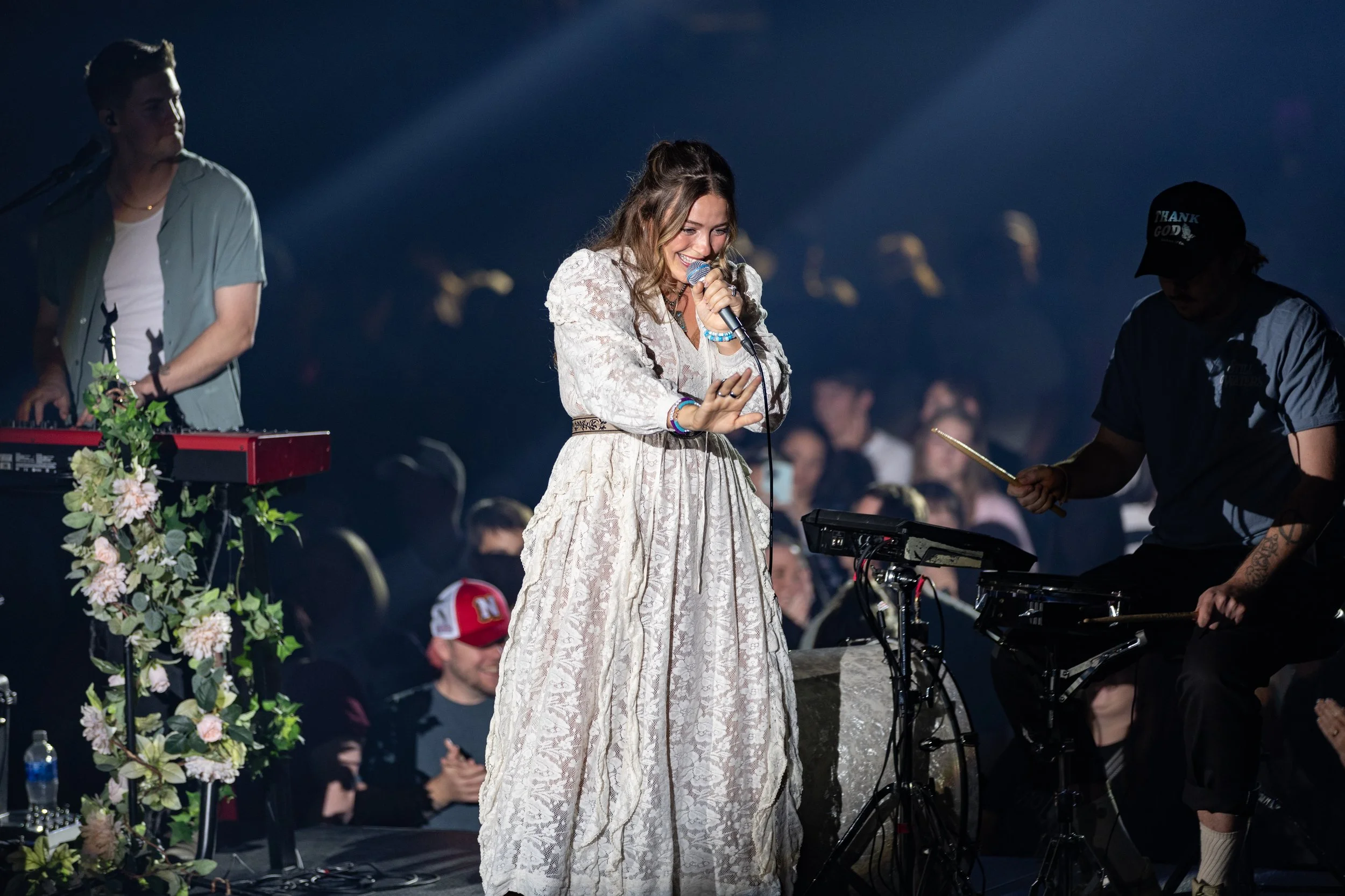 Female singer performing on stage with a keyboard and drummer, surrounded by an audience.