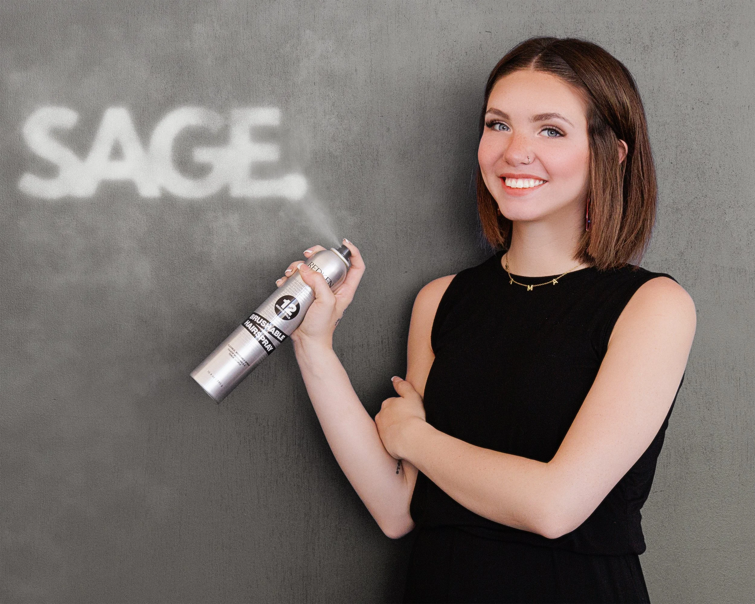 Woman smiling and holding a can of spray paint, which is creating the word 'SAGE' on a wall.