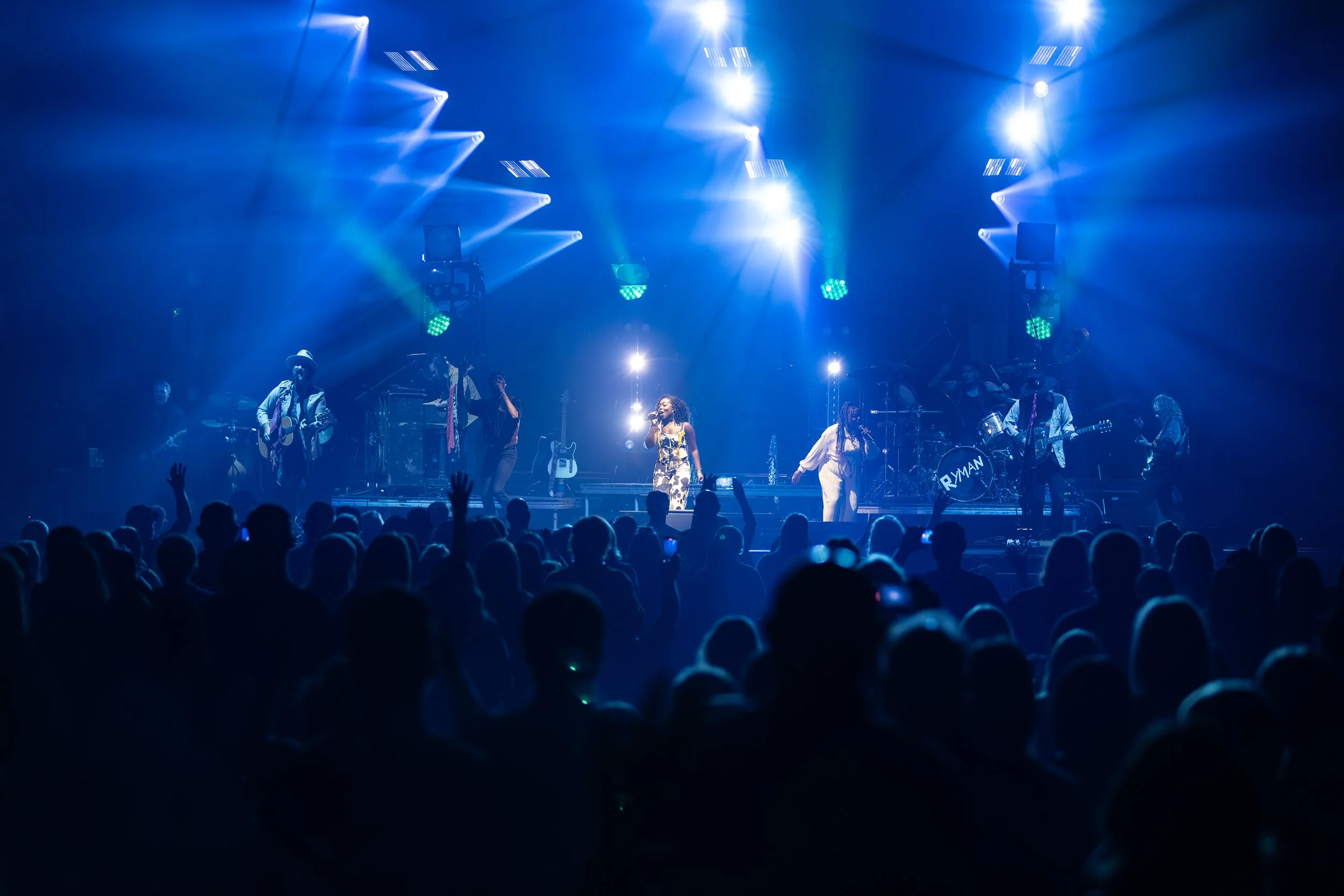 Concert stage with band performing under blue and white lights, audience with raised hands.