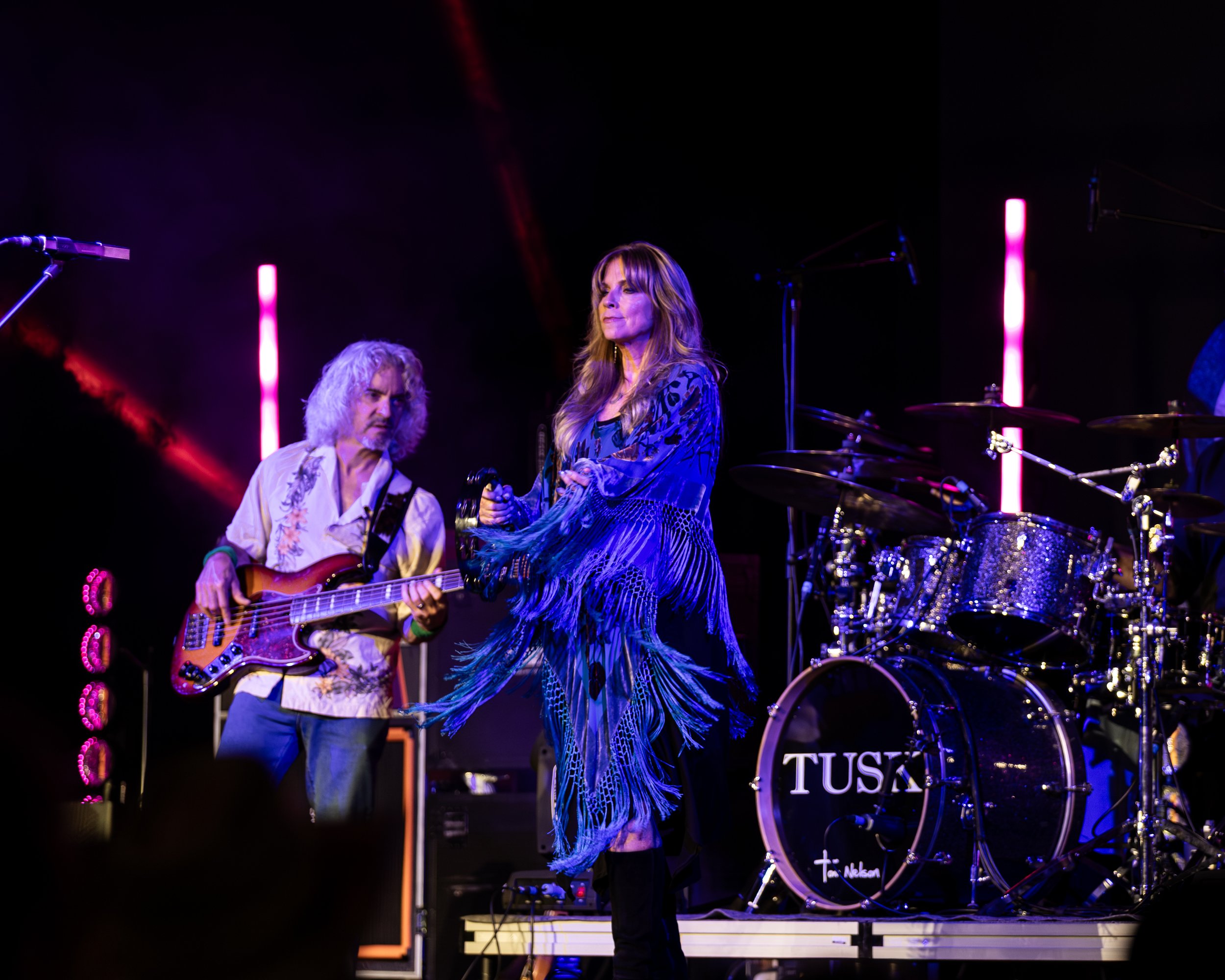 Female singer on stage with a guitarist and drum set in the background, performing at a concert.