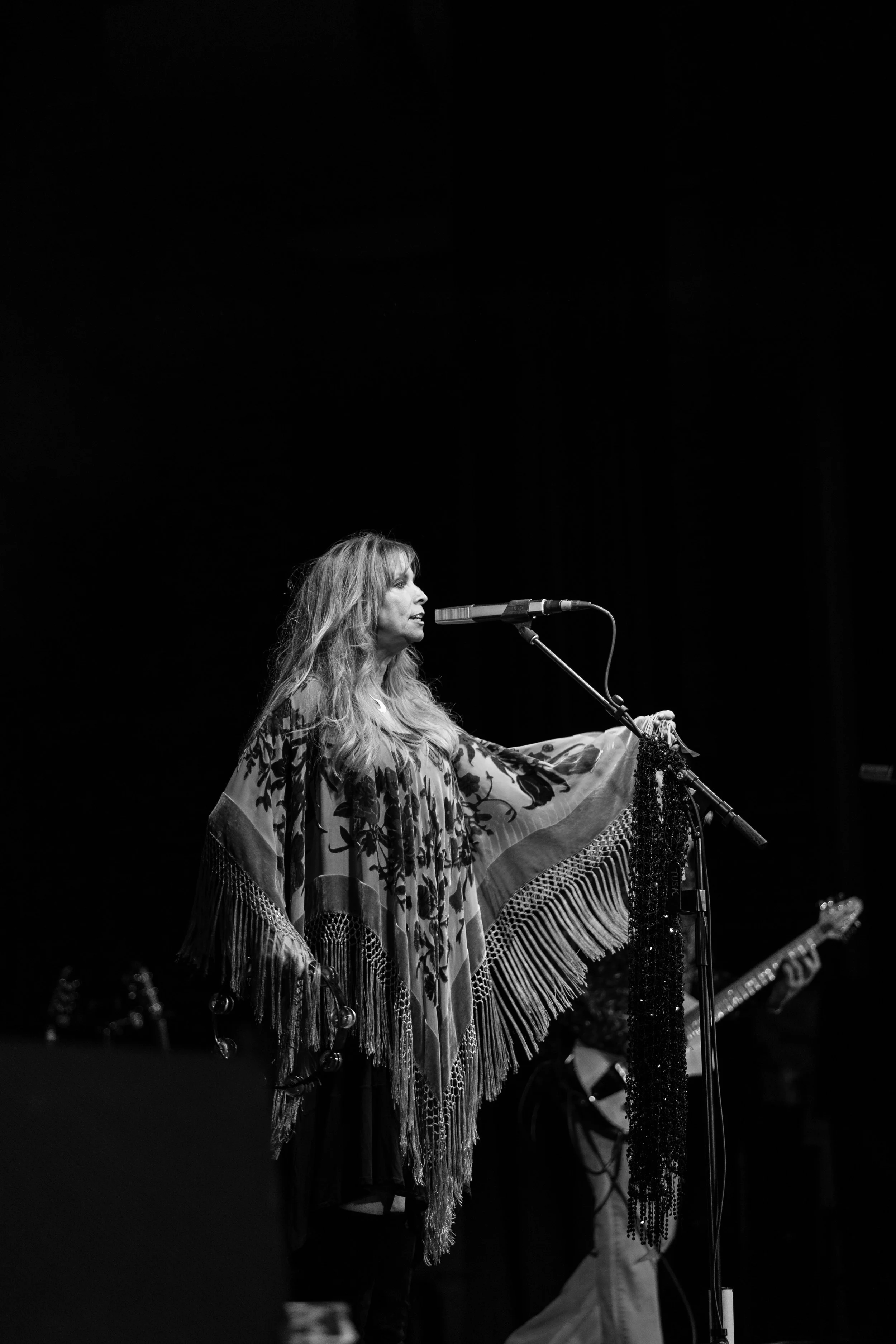 Black and white photo of a woman singing or speaking into a microphone on stage, wearing a patterned shawl with fringes, with musical instruments in the background.