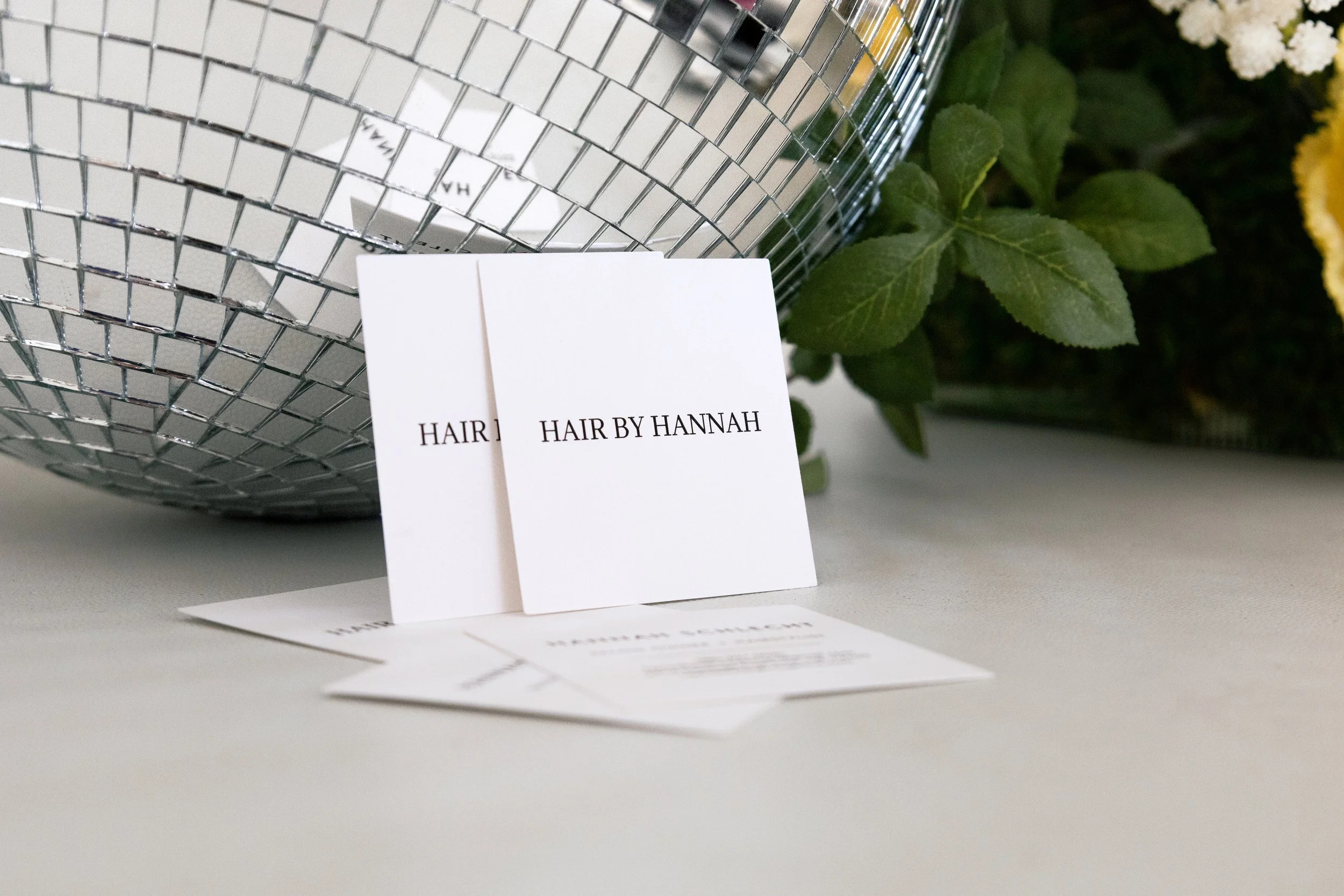 Two white cards with 'HAIR BY HANNAH' printed on them, placed on a flat surface. In the background, a silver, reflective, mosaic-patterned spherical object and some green leaves are visible.