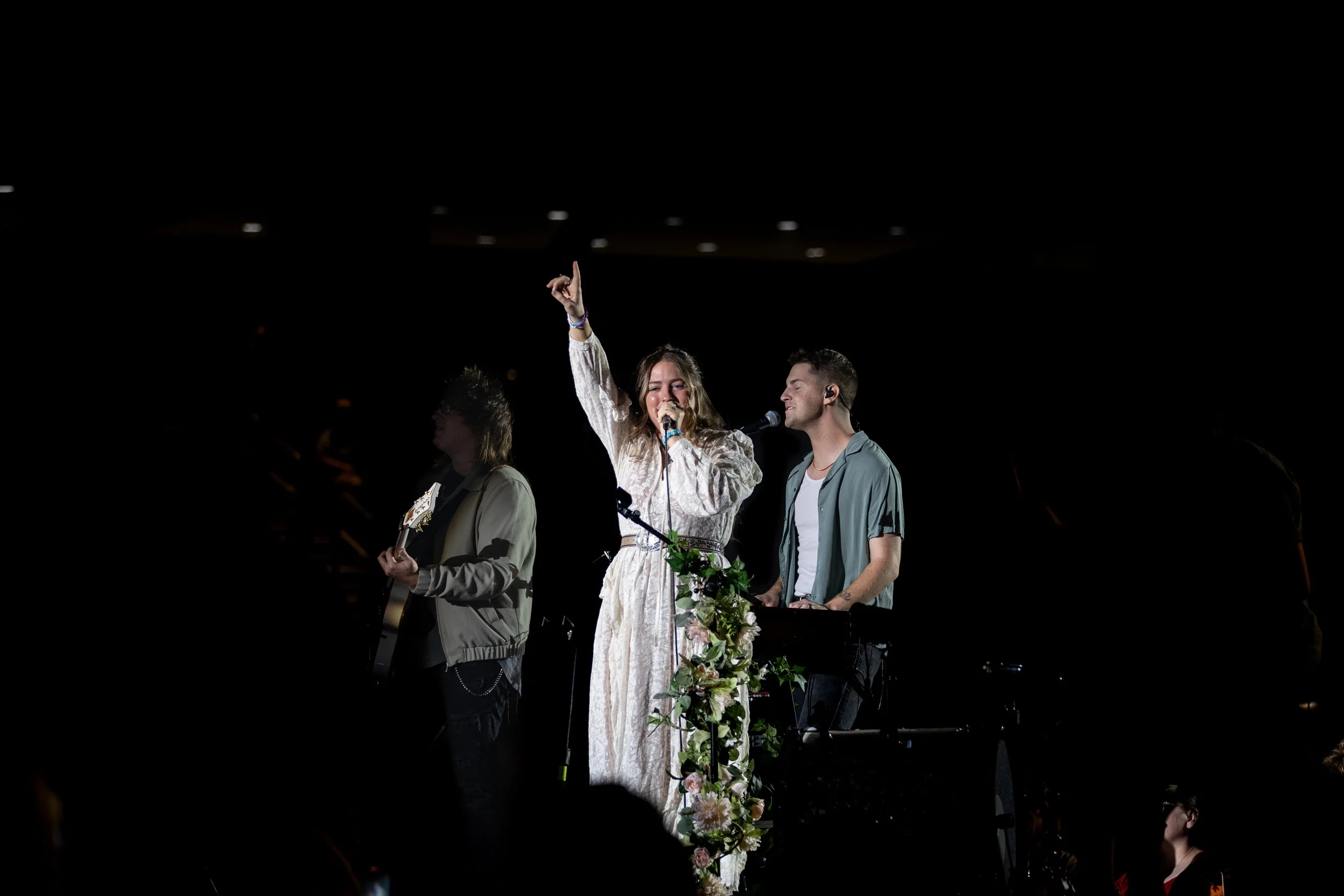 A woman singing into a microphone and raising her hand, flanked by a man playing keyboard and another person playing guitar, on a stage decorated with flowers.