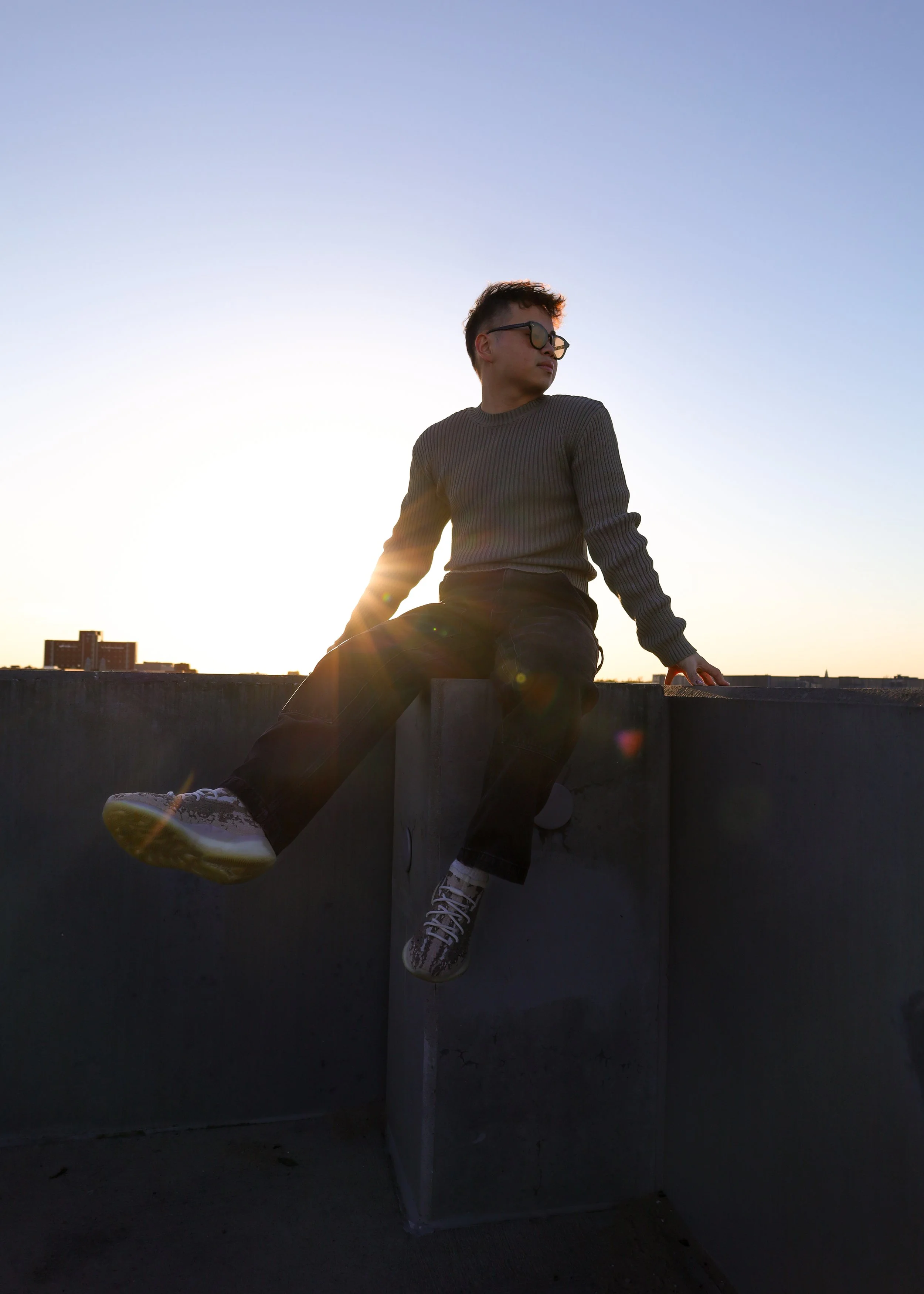A person sitting on the edge of a rooftop at sunset, wearing glasses, a gray sweater, black pants, and patterned sneakers, with the city skyline in the background.