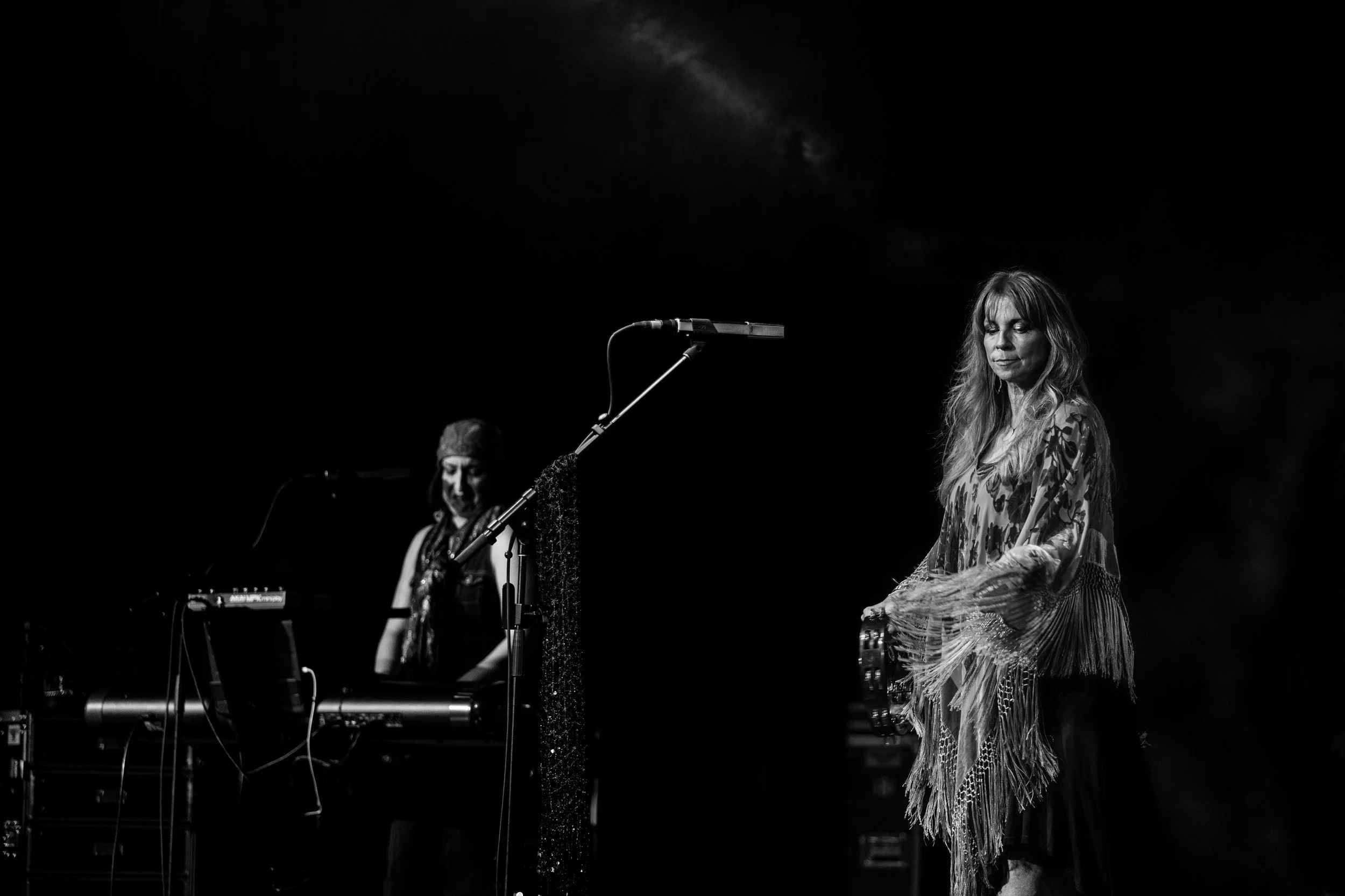 Black and white photo of two women on stage, one playing a tambourine with eyes closed, the other standing with eyes closed, wearing a fringed dress, in a dark setting.