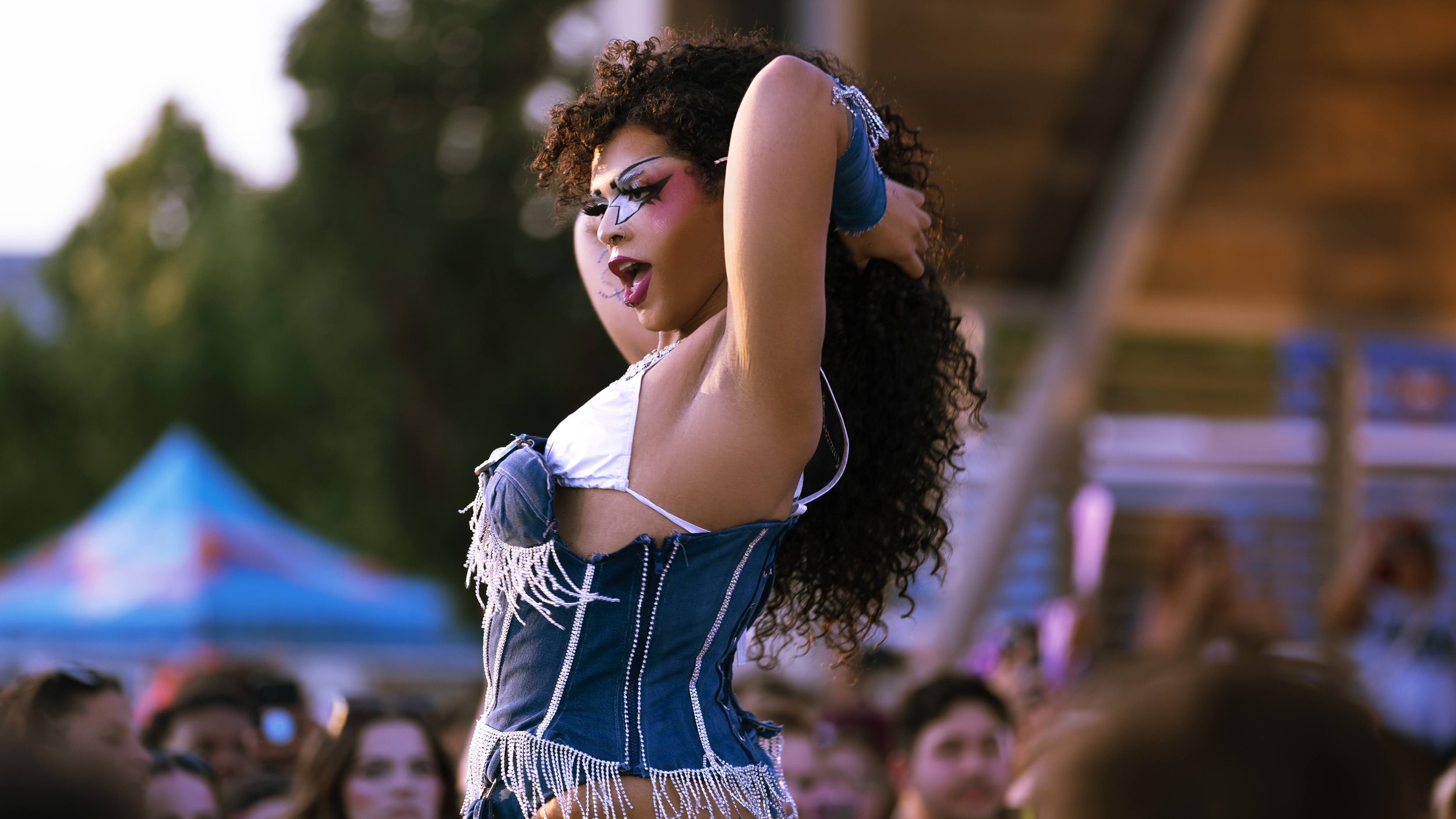 Performer with curly hair and bold makeup dancing at an outdoor event, wearing a decorated outfit with fringe.