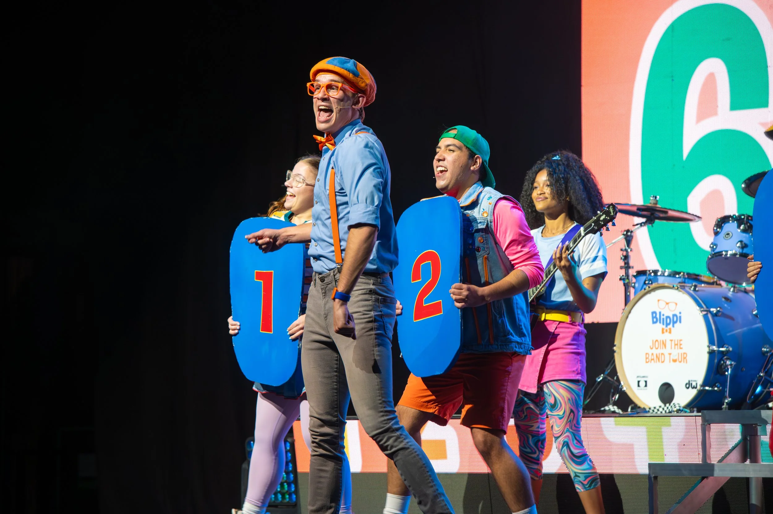Children and an adult perform on stage with large colorful signs indicating numbers, a drum set with a sign reading 'Join the Band Tour', and a bright, playful backdrop.
