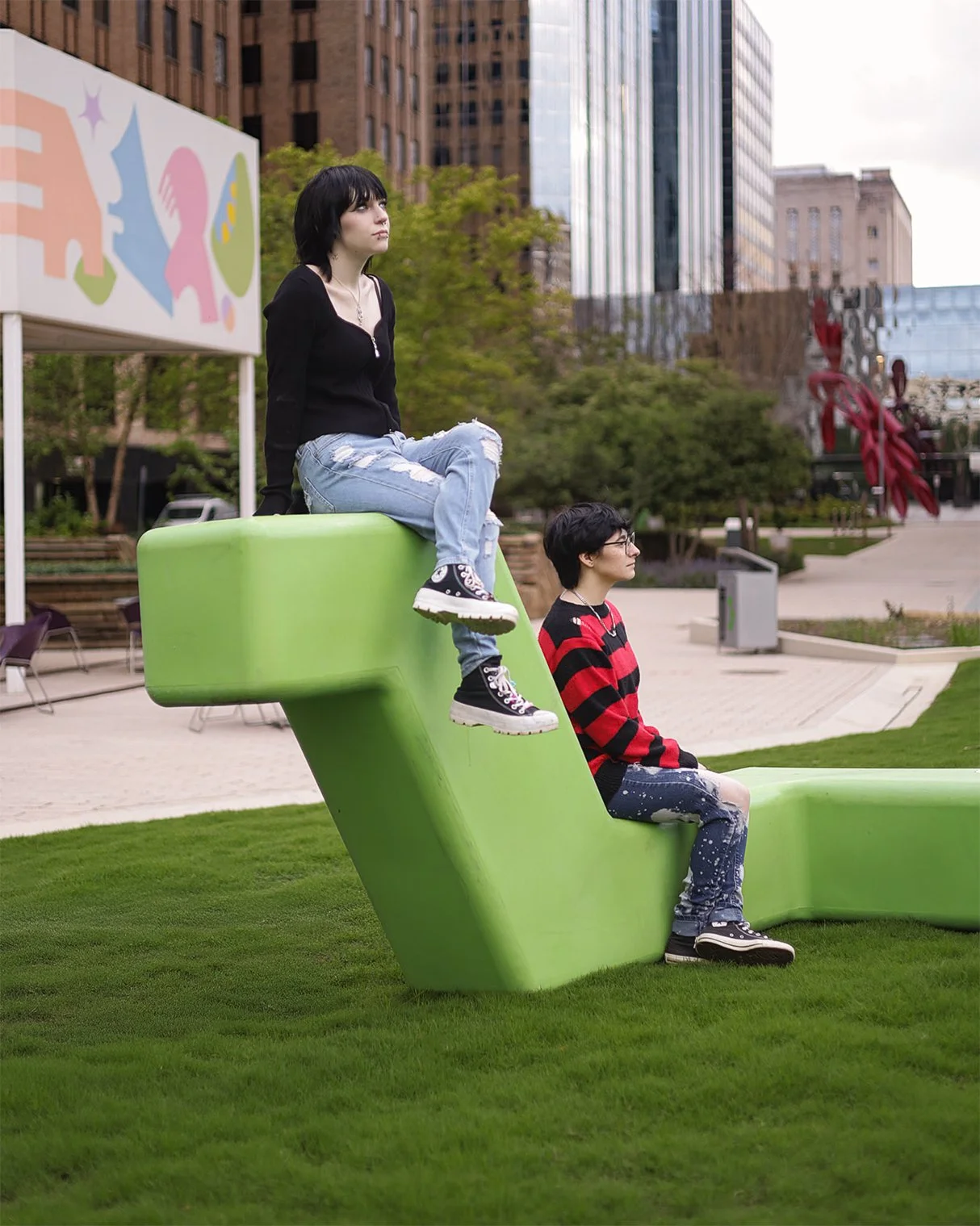 Two young women with short black hair, one sitting on a large green sculpture and the other sitting on the ground, in an urban park with tall buildings and trees in the background.