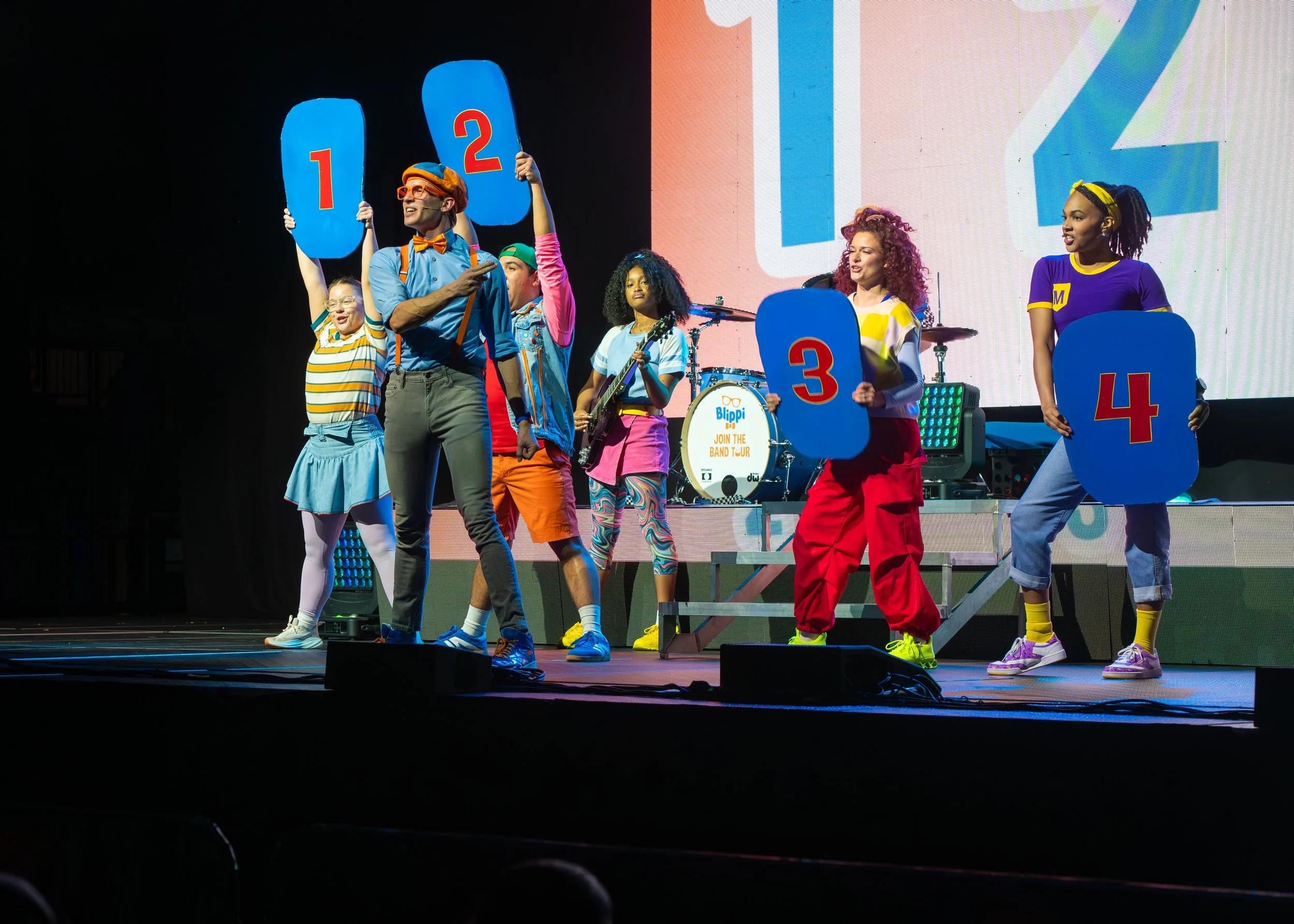 A group of performers dressed in colorful costumes on stage, holding numbered signs from 1 to 4, participating in a lively show with a drum set in the background.