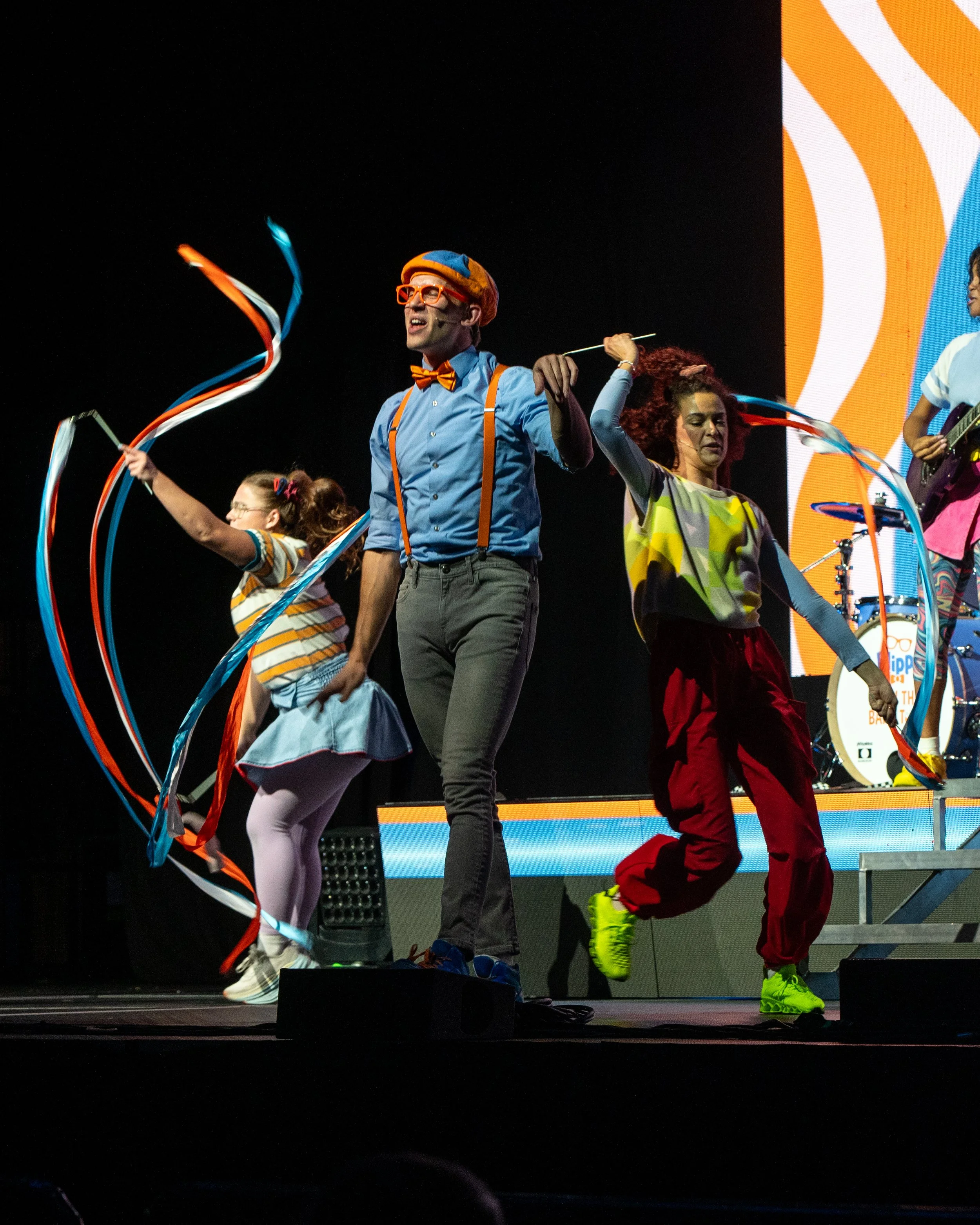 A group of performers on stage dancing with colorful ribbons, dressed in bright, playful costumes.