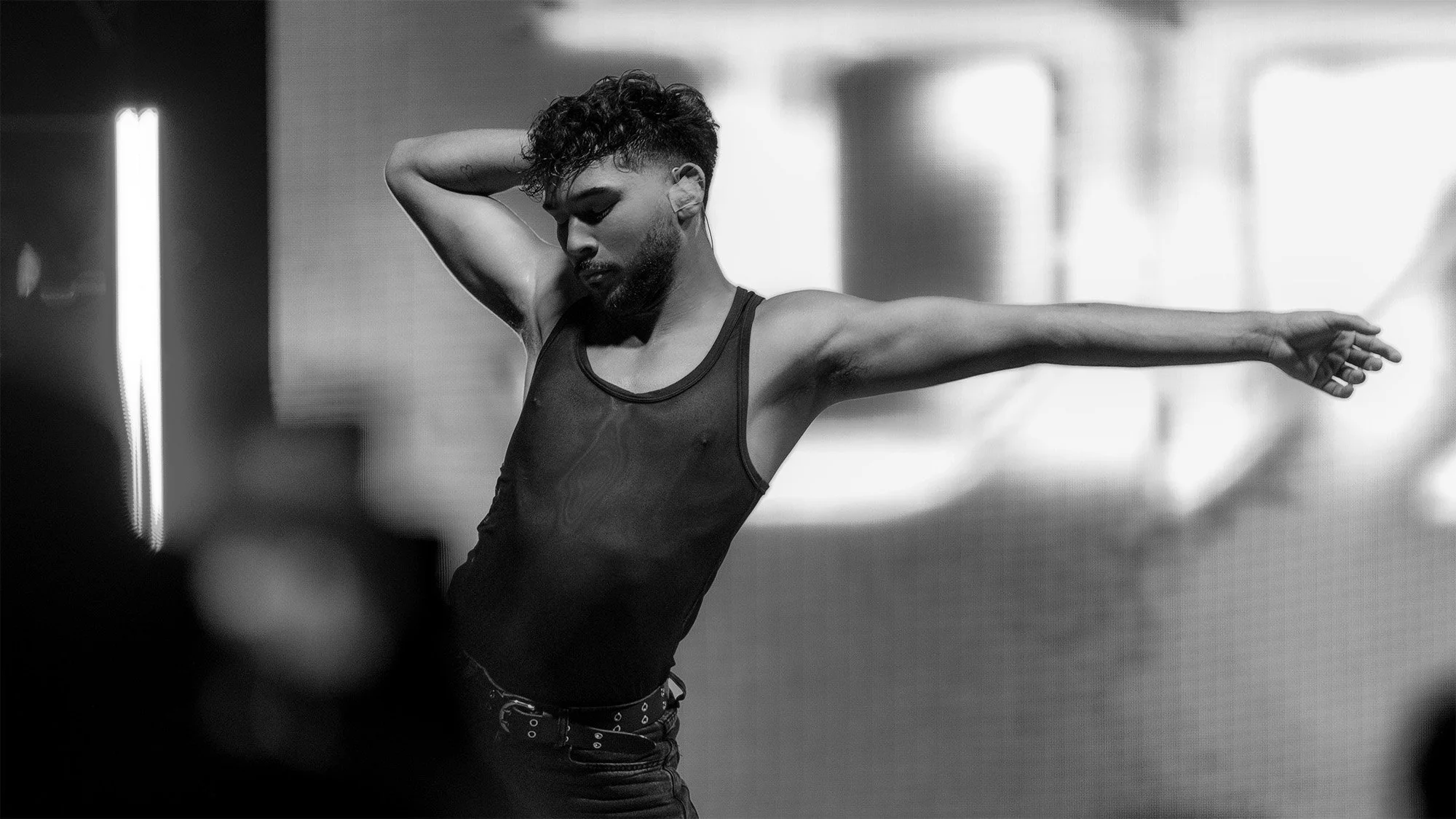 A black and white photo of a young man with curly hair and a beard, stretching with one arm behind his head and the other arm extended outward in a dance or exercise pose.