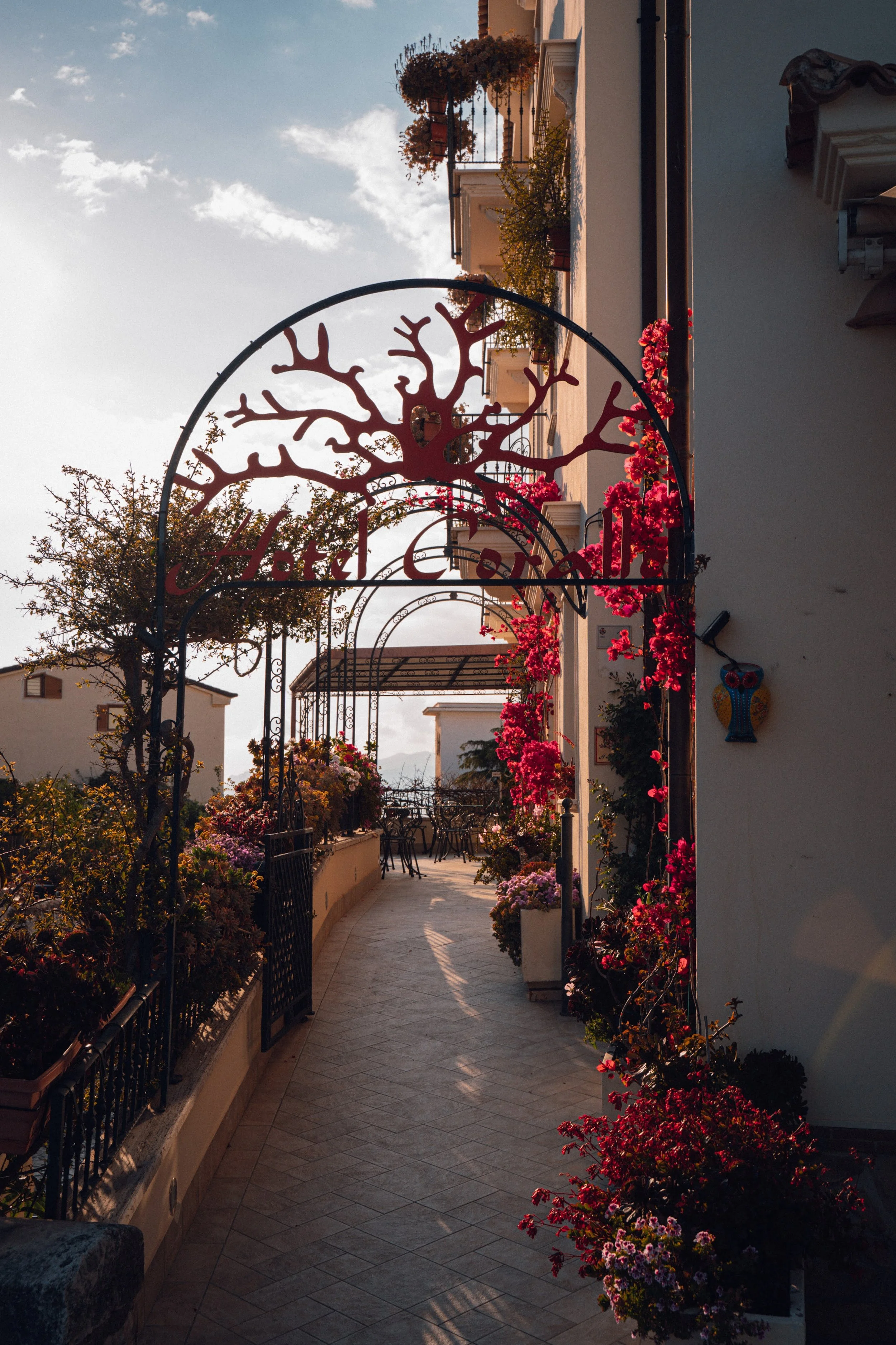 A balcony with a curved gate and a sign that reads "Hotel". The balcony is decorated with pink and purple flowers and has seating. The scene is illuminated by late afternoon sunlight, with buildings and trees in the background.