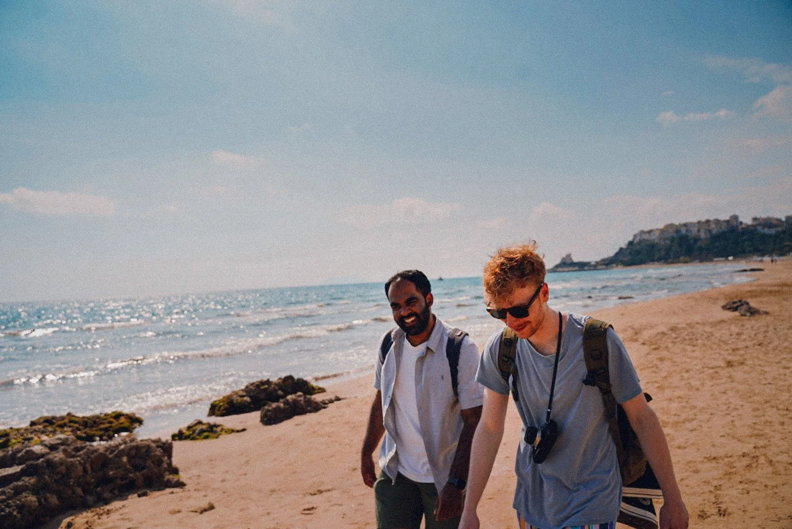 Two young men walking on a sandy beach with rocks, ocean waves, and a distant headland under a blue sky.