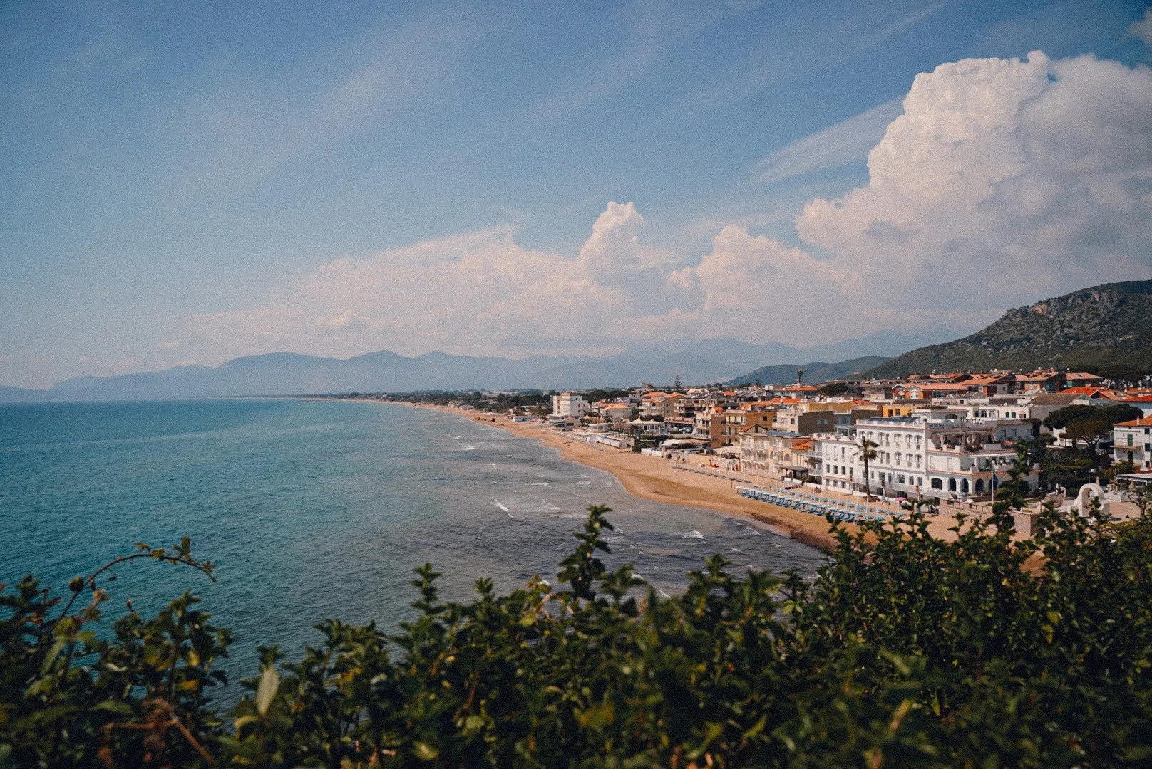 A coastal town with buildings along a sandy beach, overlooking the ocean, with distant mountains and partly cloudy sky in the background.