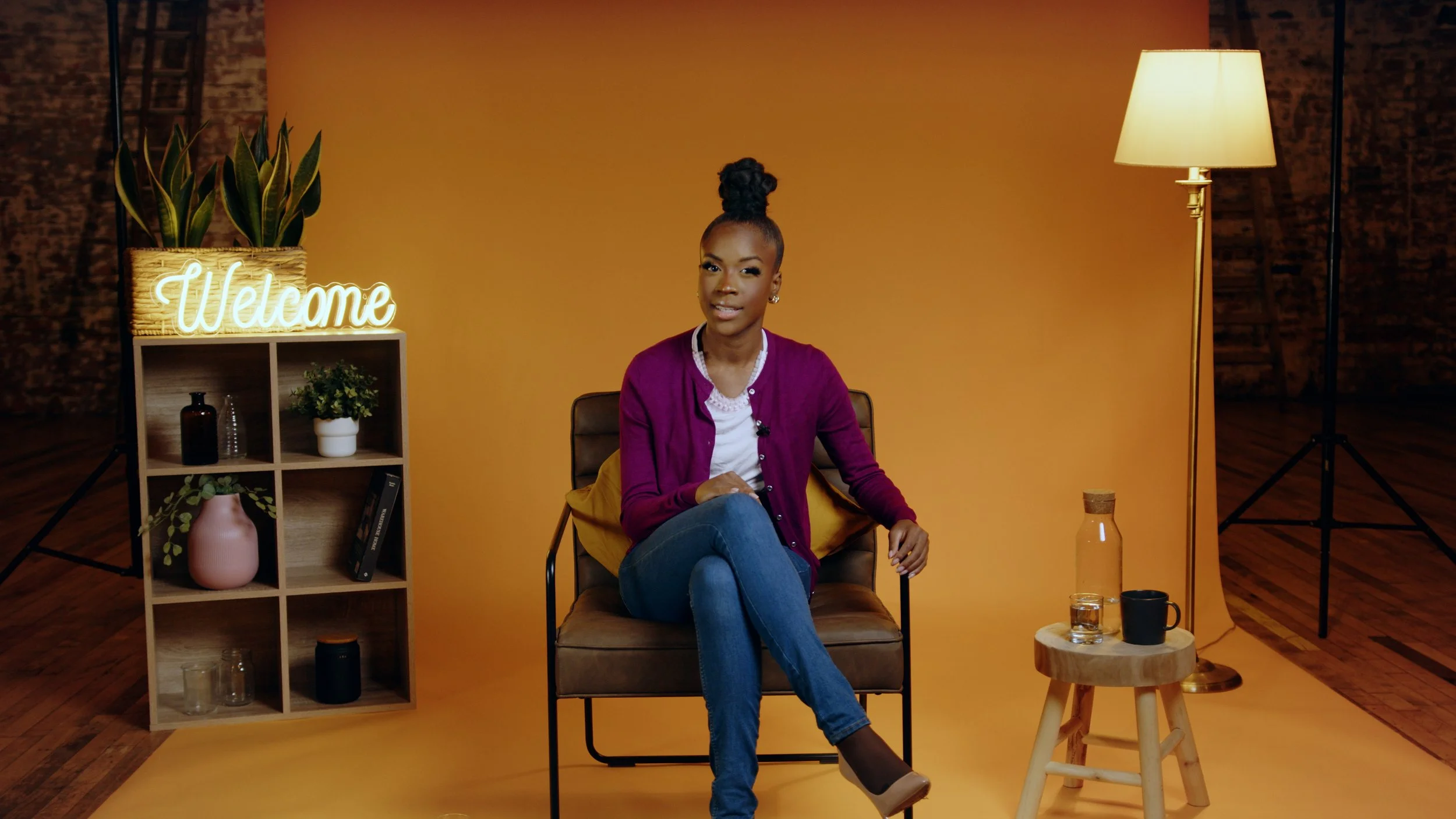 A woman sitting on a brown chair with crossed legs, wearing a white shirt and a purple cardigan, in a studio with yellow backdrop. To her left, a wooden shelf displaying plants and decorative items with a neon