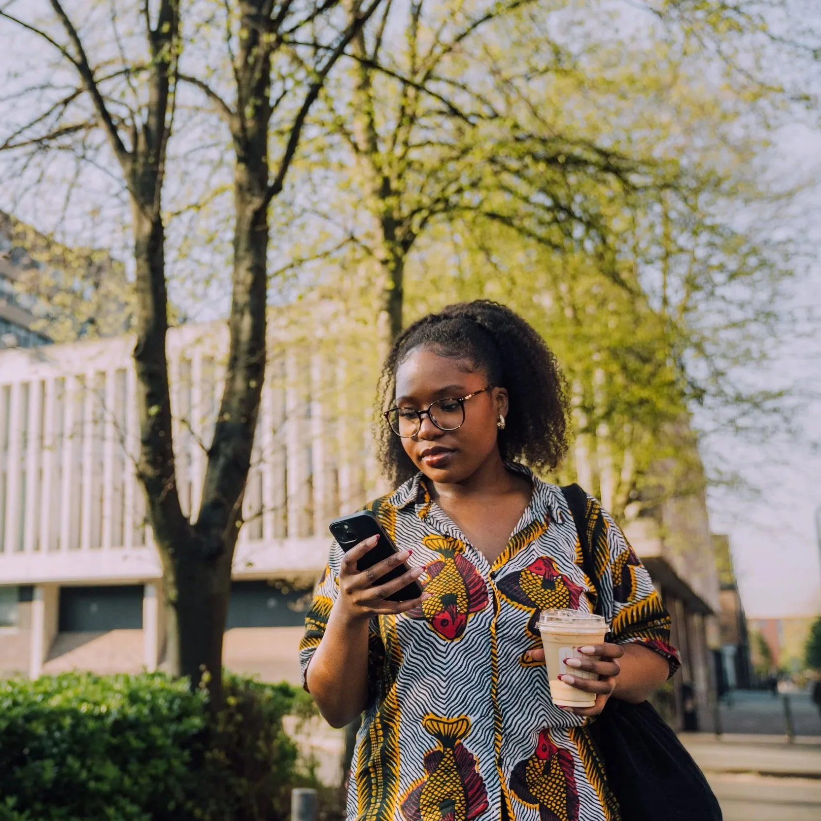 Young woman with glasses and curly hair looking at her phone while holding a coffee cup, outdoors with trees and buildings in the background.