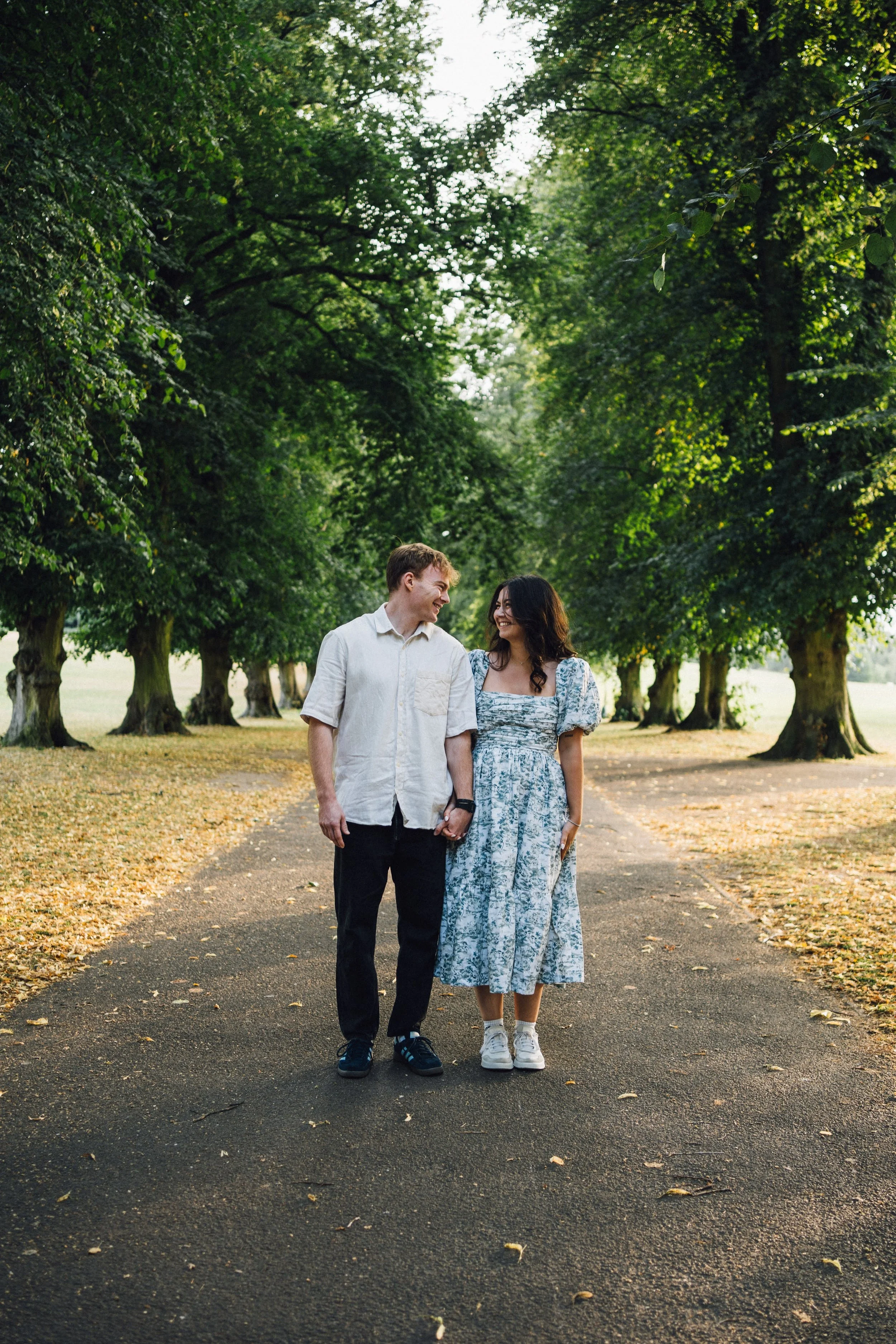 A young couple walking hand in hand on a tree-lined path, smiling at each other.