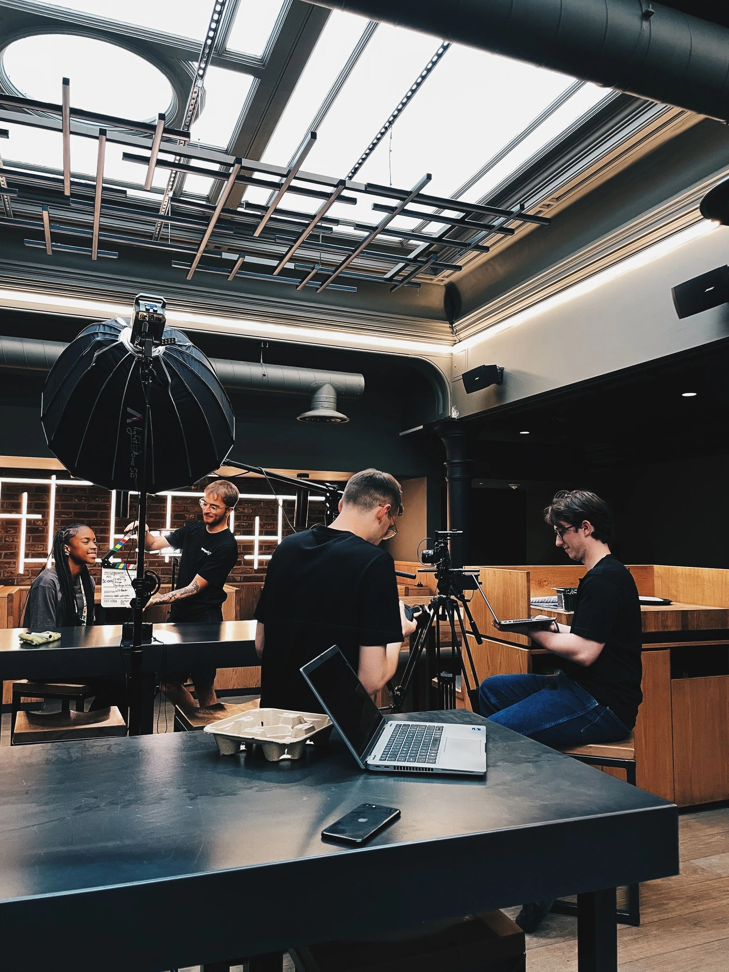 A video production crew filming a woman with braids in a modern indoor space with large skylights, professional lighting equipment, a camera on a tripod, and a laptop on the table.