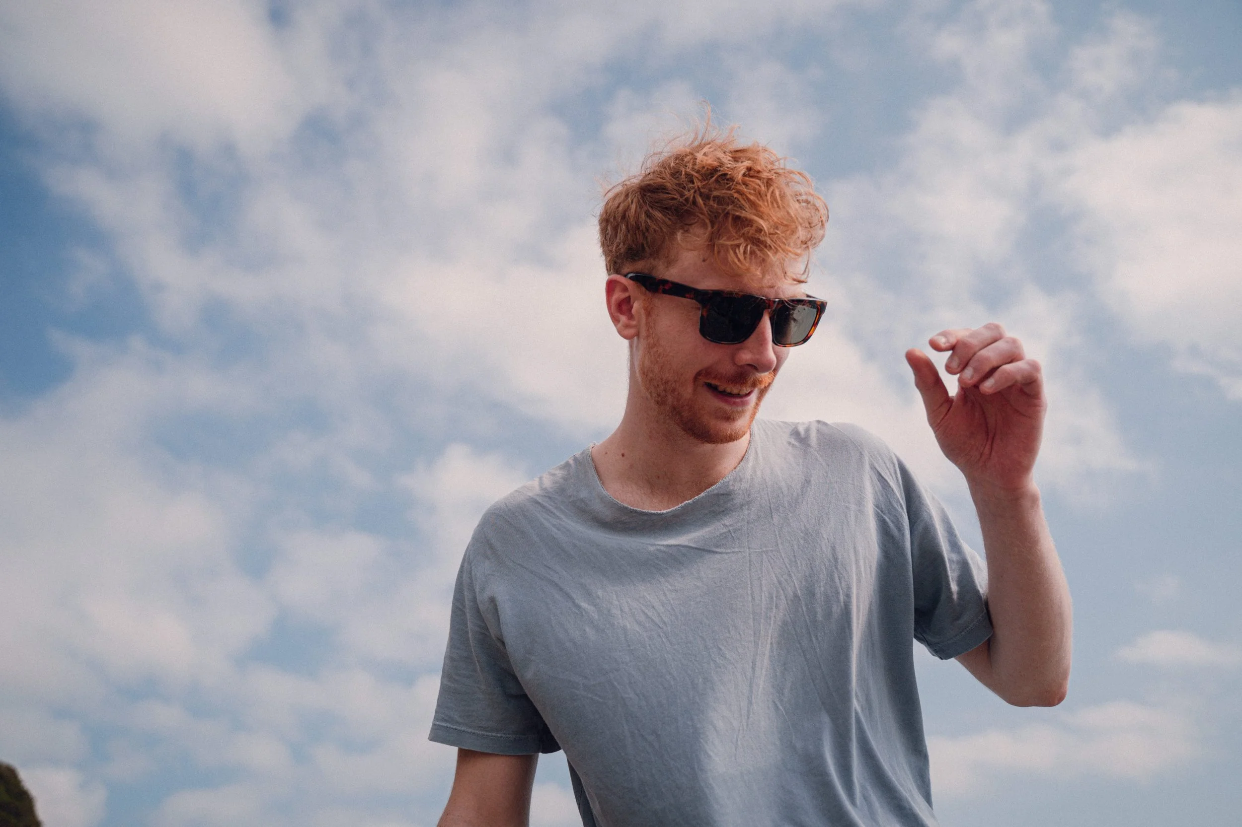 Young man with curly red hair and sunglasses smiling outdoors against blue sky with clouds, wearing a light gray t-shirt.