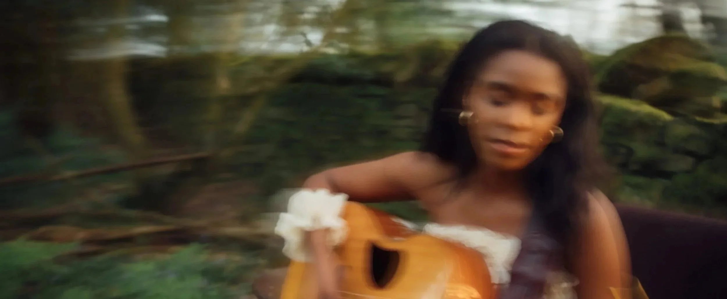A woman with long dark hair playing an acoustic guitar outdoors among green trees, slightly blurred due to motion.