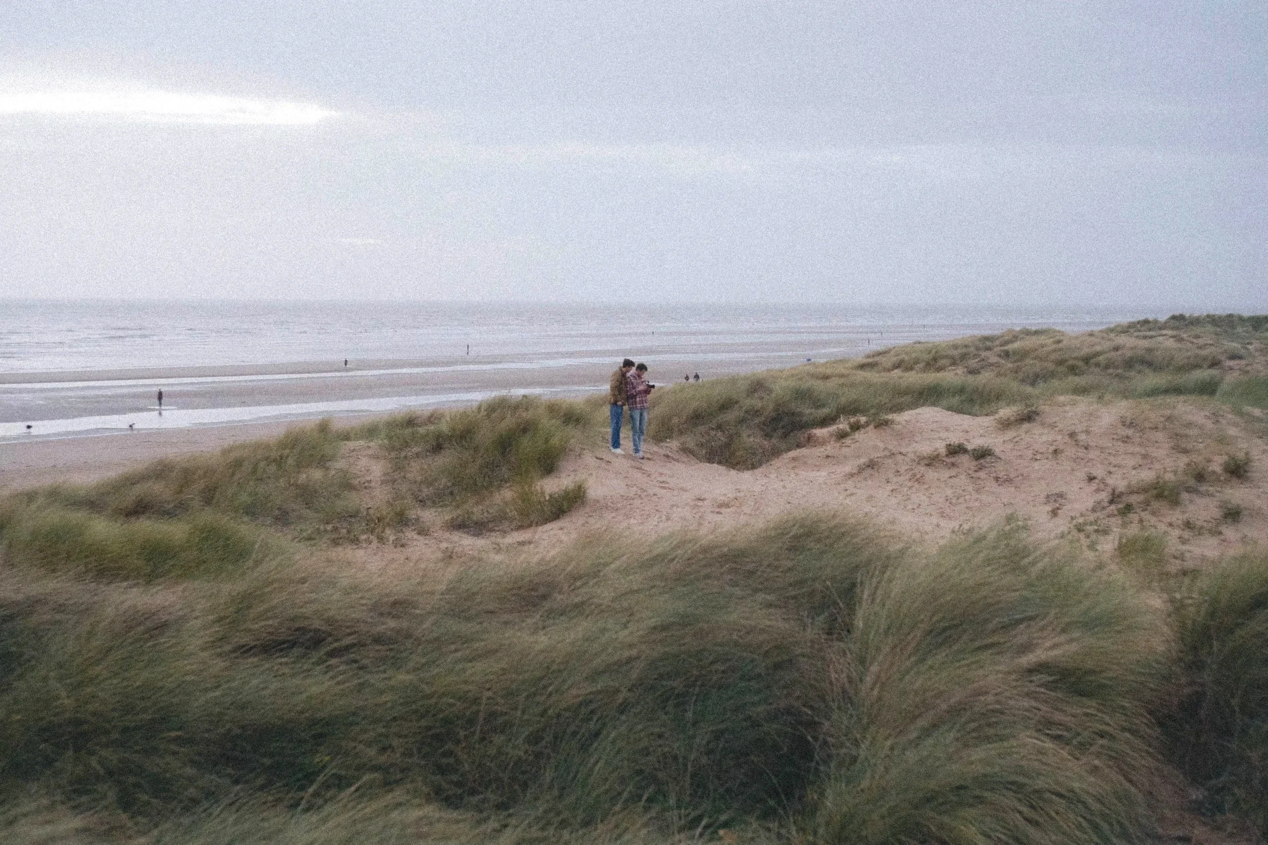 Two people stand on sand dunes overlooking a beach with gentle waves under a cloudy sky, while a few people walk along the shoreline.