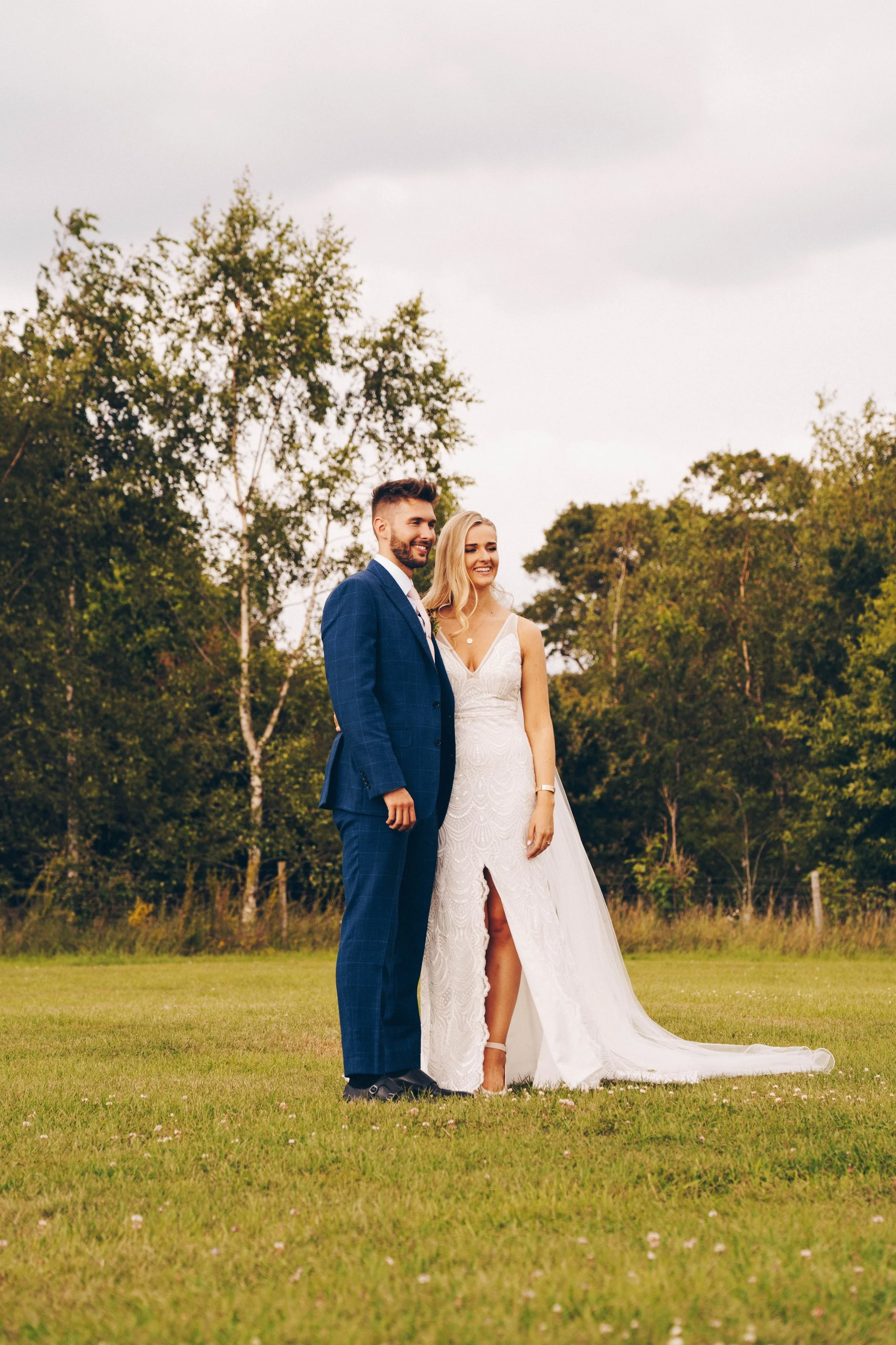 A happy bride and groom standing together on a grassy field with trees in the background, smiling outdoors.