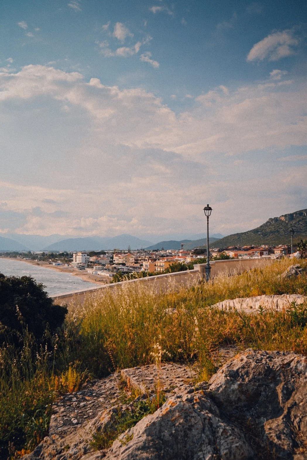 A scenic view of a coastal town with a beach, buildings, and mountains in the background, with a lamp post on a grassy and rocky foreground.
