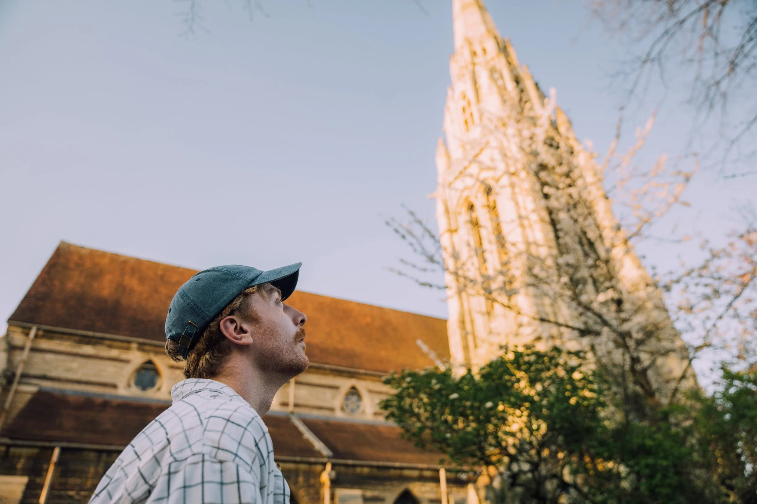 A young man wearing a cap and a checkered shirt looking up at a tall church steeple during daytime.