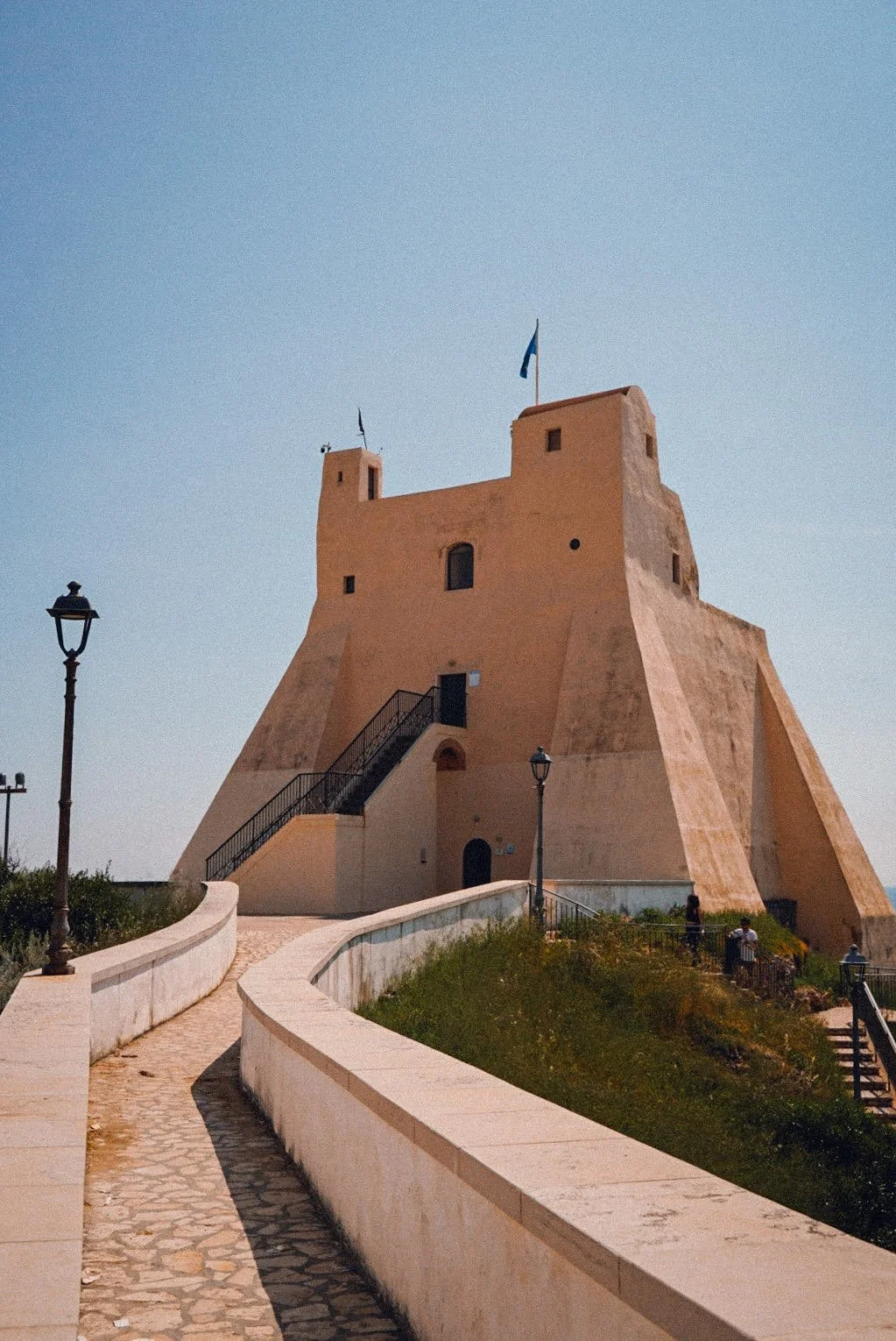 A large beige fortress with flags on top, situated on a small hill. A cobblestone path and lampposts lead up to it, with some people nearby enjoying the sunny day. Clear blue sky in the background.