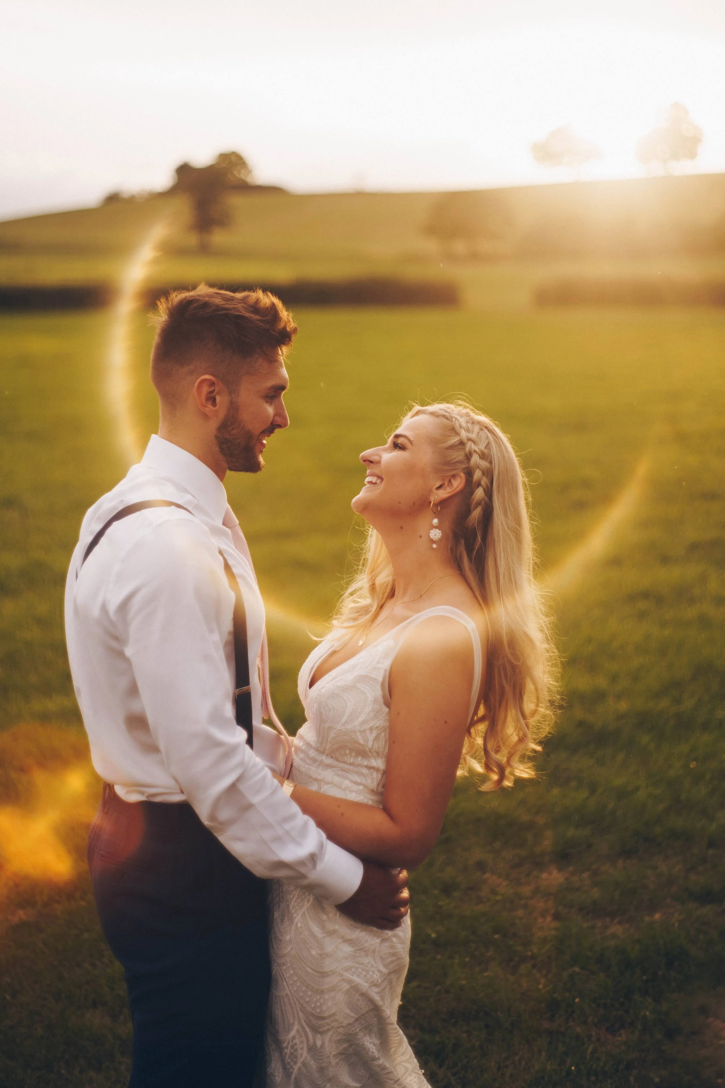 A couple is standing outdoors, gazing into each other's eyes with smiles, during sunset in a green field, with a bright, glowing aura surrounding them.