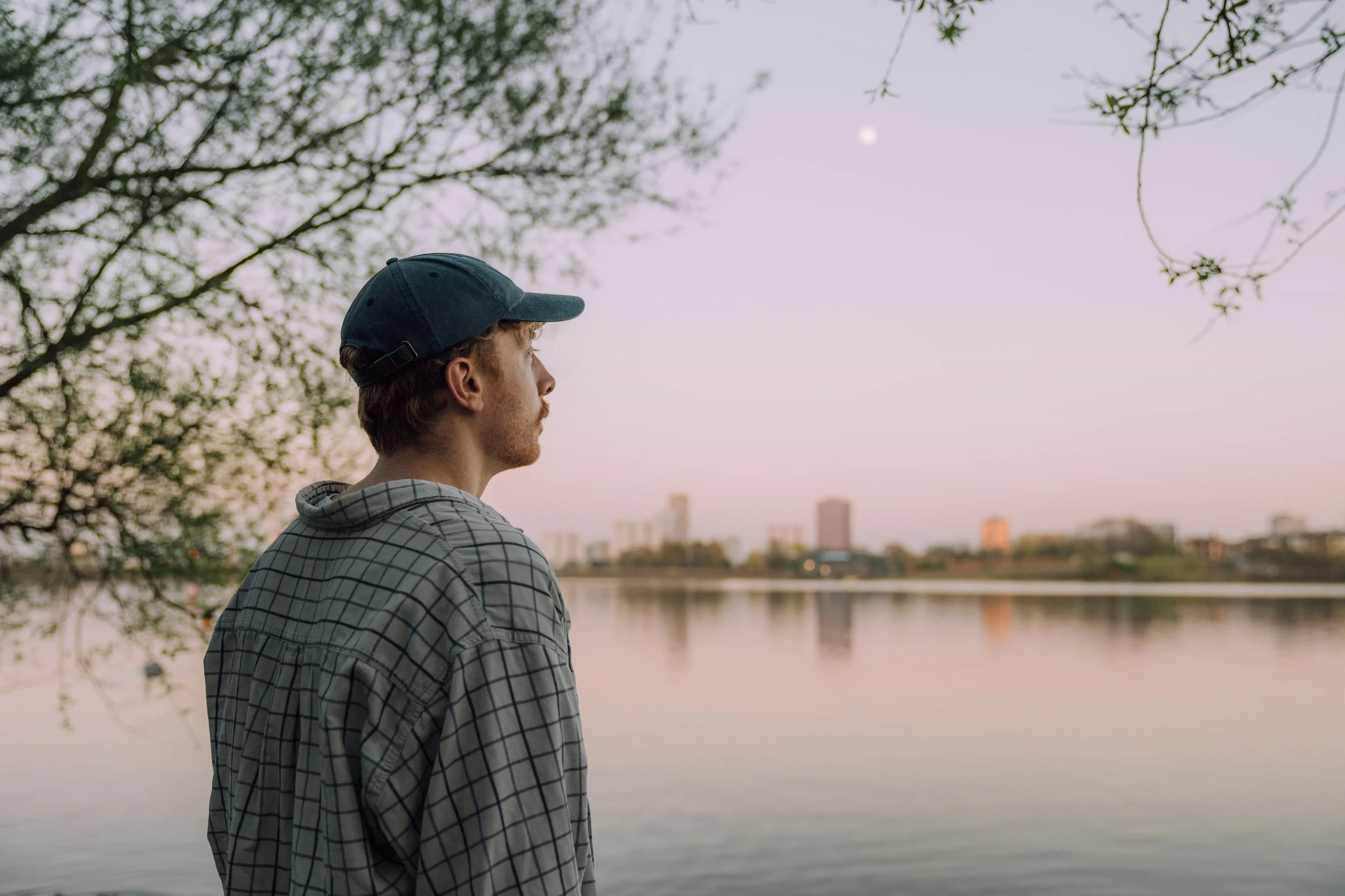 A young man wearing a cap and checkered hoodie standing by a body of water, looking at the city skyline with buildings, during dusk or dawn.