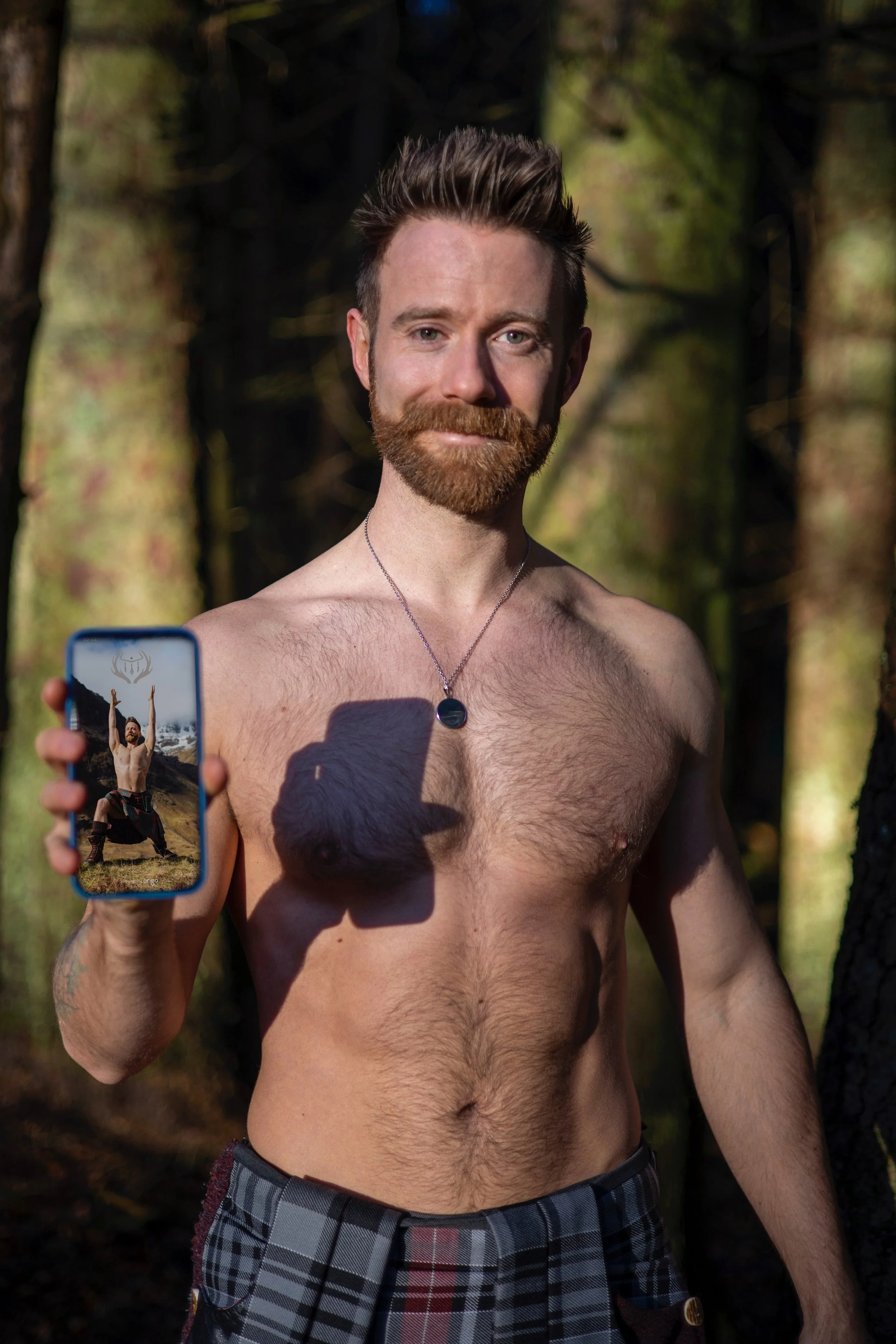 A shirtless man with a beard holding a phone showing himself in a different pose in a forested area.