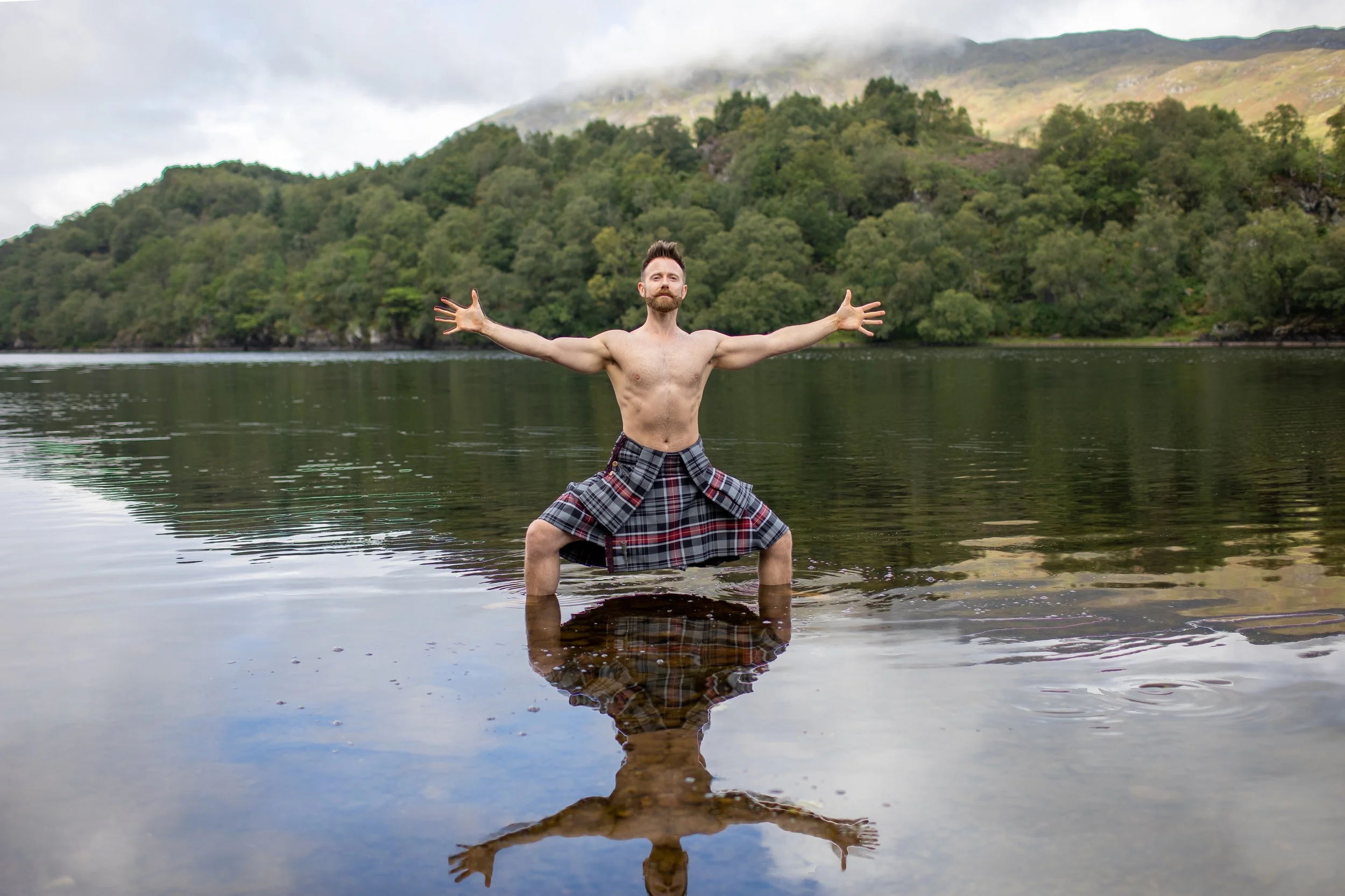An image of a man in a kilt doing a yoga pose called horse while standing in a loch.