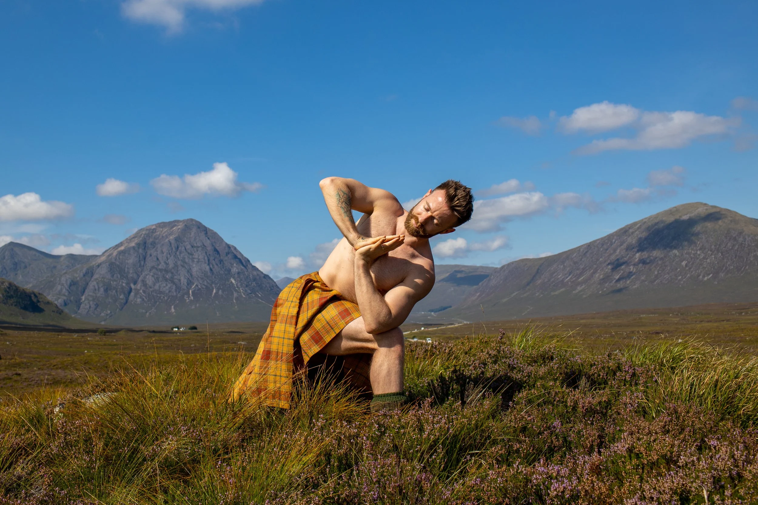 A shirtless man with a plaid kilt practicing yoga outdoors in a grassy field with mountains in the background under a partly cloudy sky.