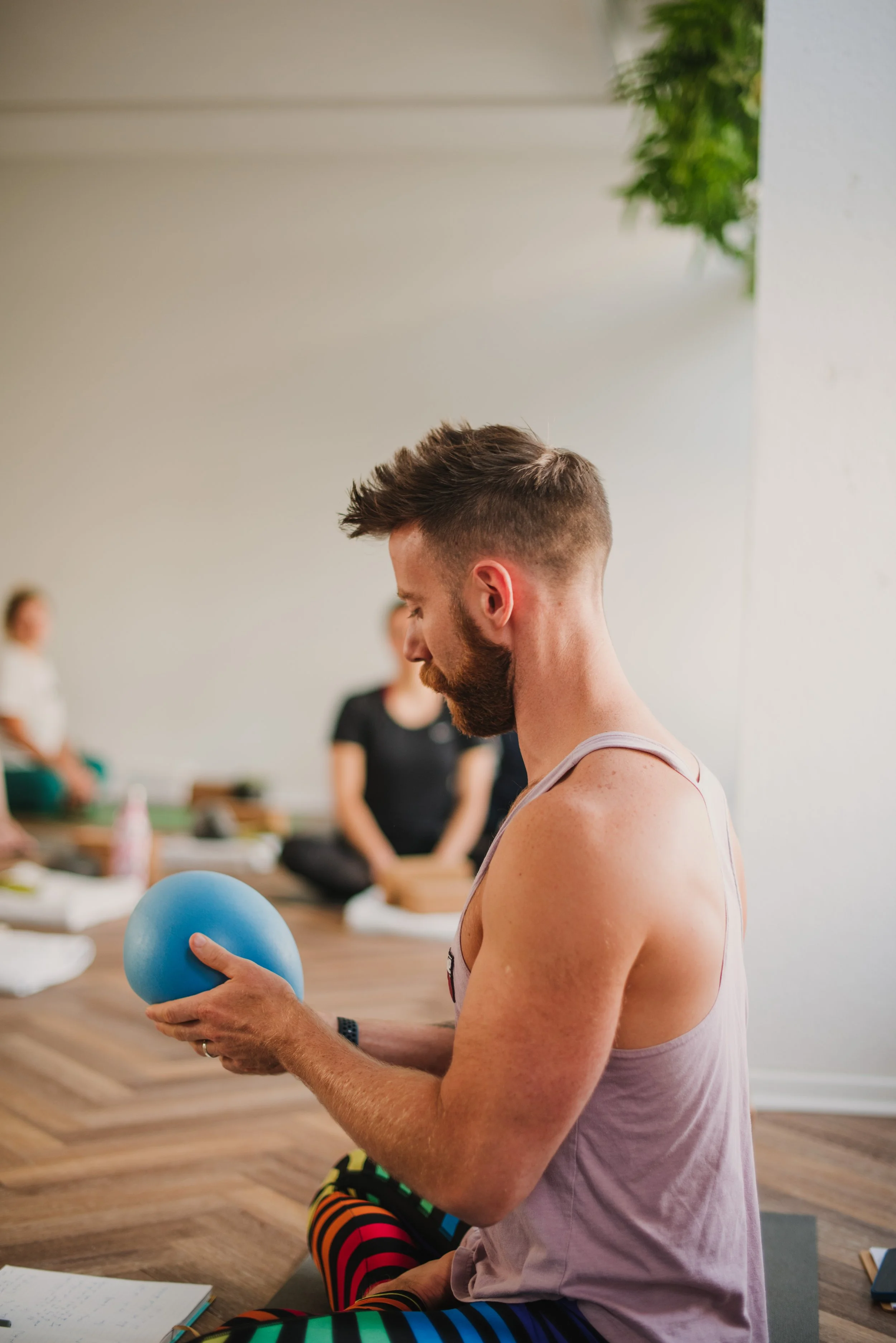 Man with a beard and colorful striped pants sitting on the floor, holding a blue exercise ball, with people in yoga poses in the background during a yoga class.