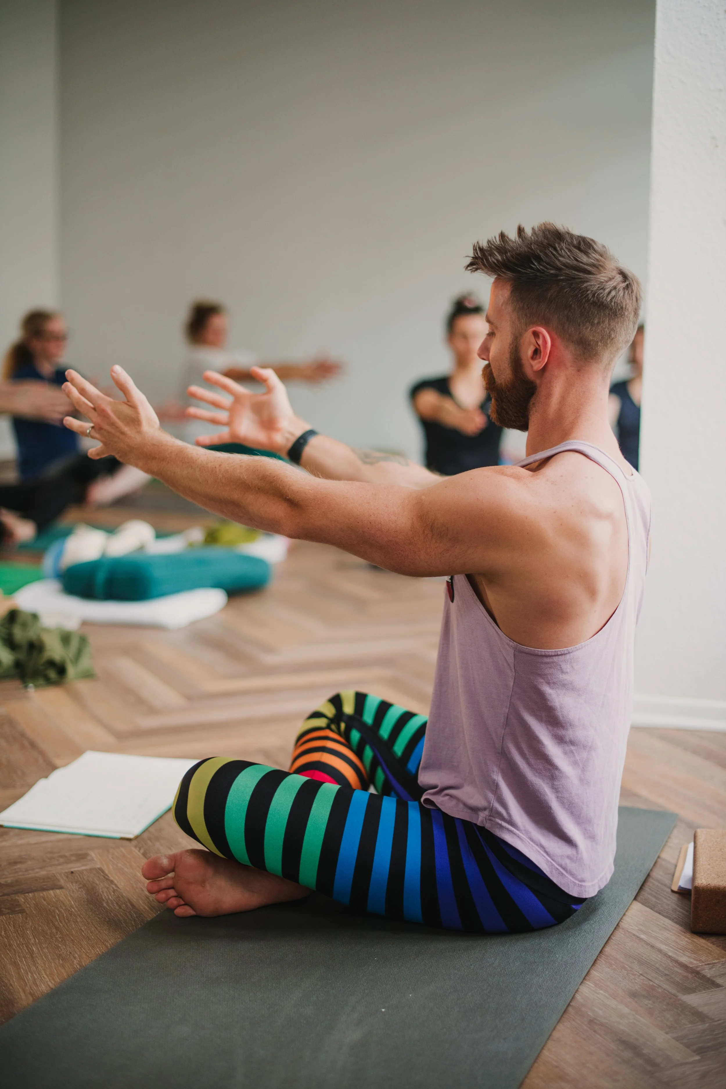 Man in a yoga class sitting cross-legged on a yoga mat, leading the group in a stretching exercise with others in the background.