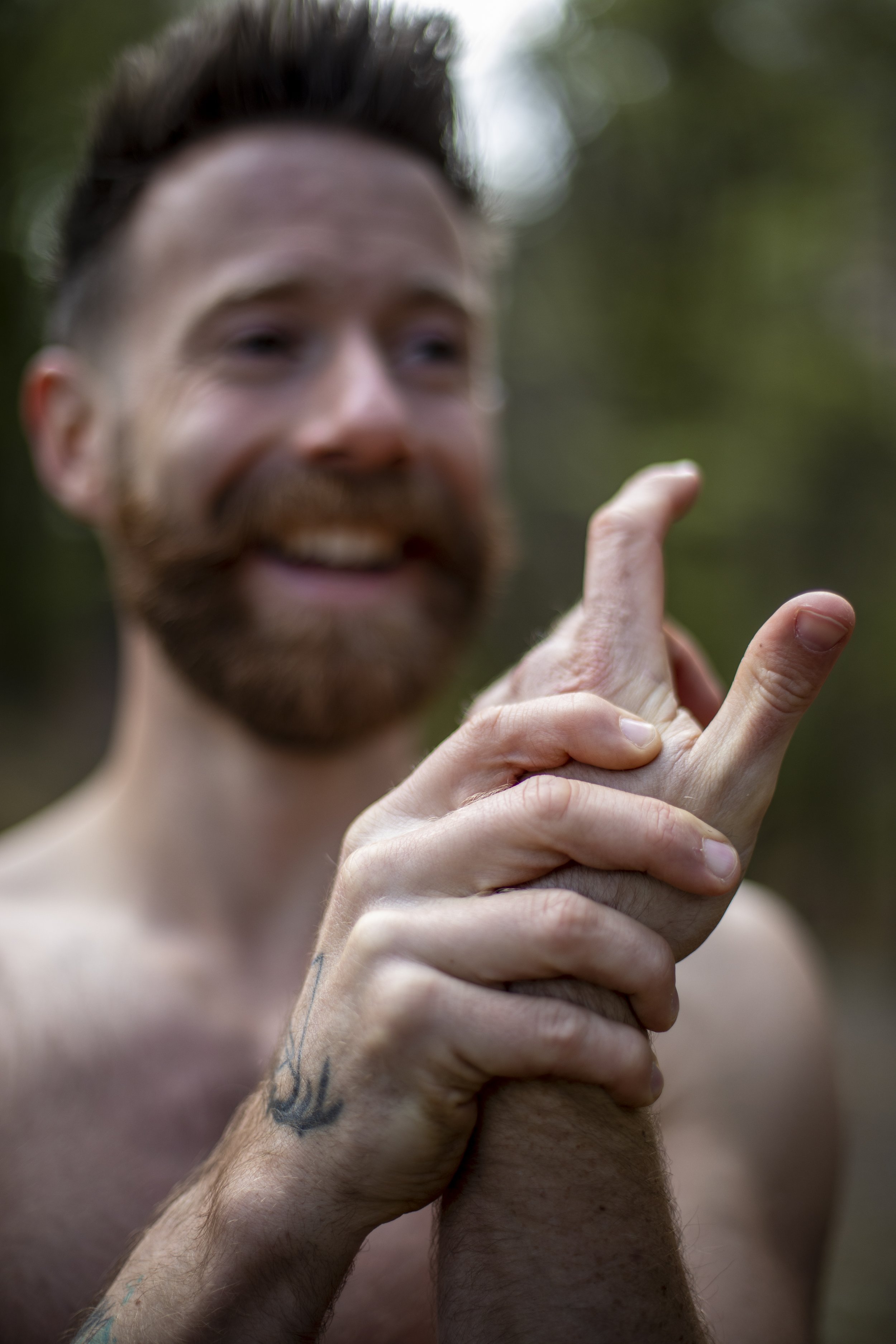 A shirtless man with a beard smiling and making a playful finger gun gesture with his hand, with a blurred outdoor background.
