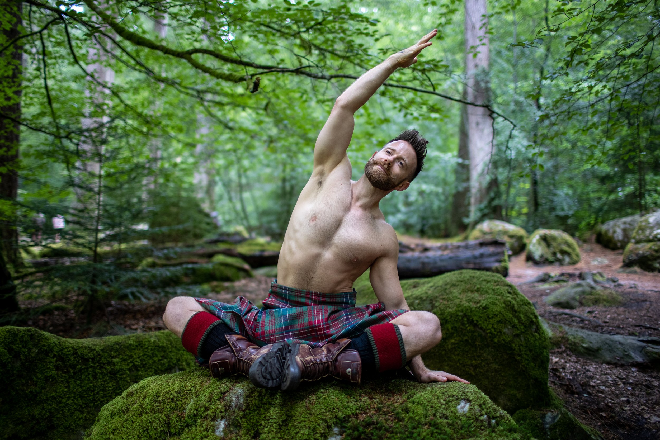 A shirtless man with a beard and plaid kilt performing a yoga pose on moss-covered rocks in a forest.
