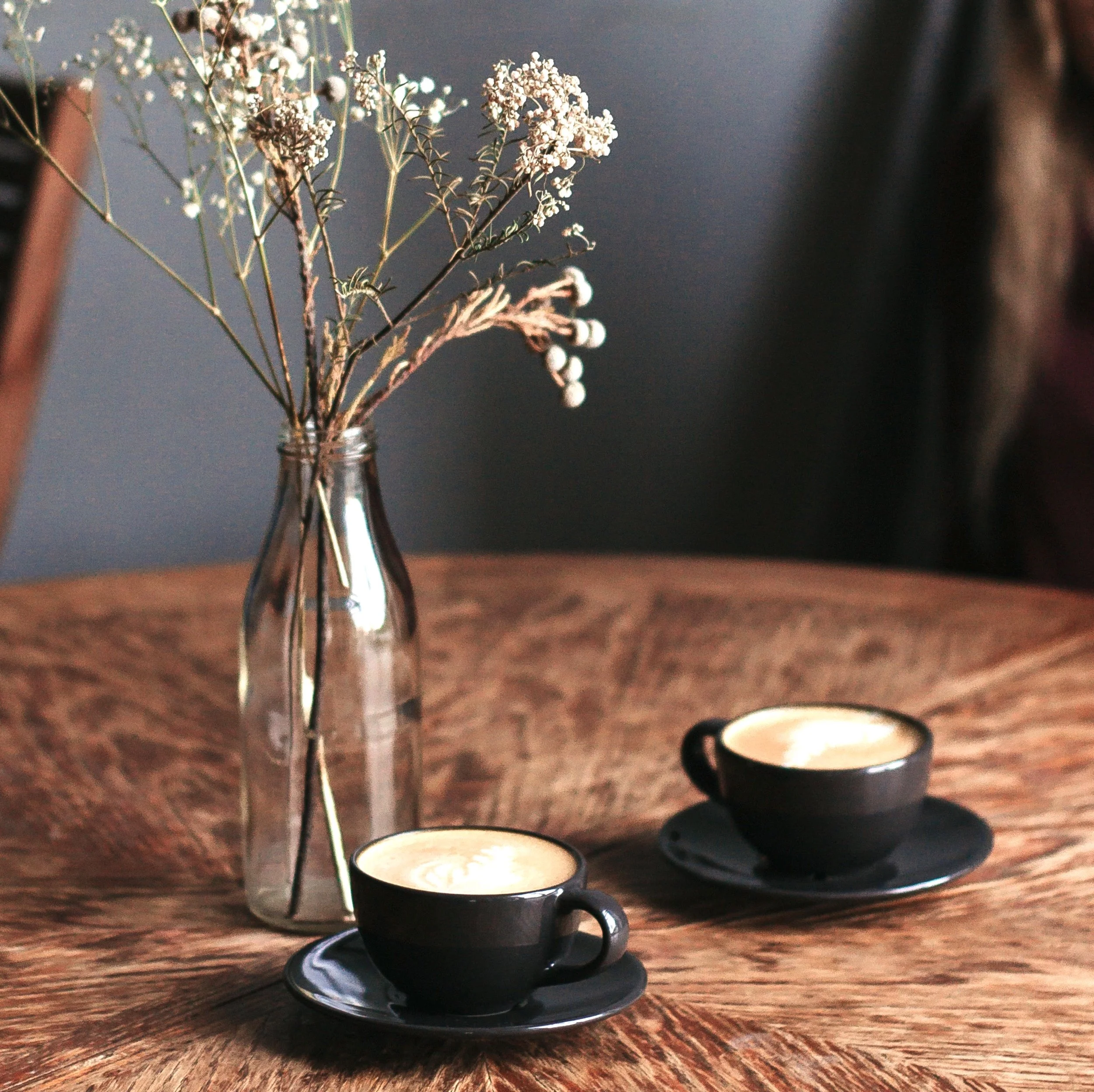 A wooden table with a glass vase containing dried white flowers and two black cups filled with a frothy beverage, each on a matching black saucer.