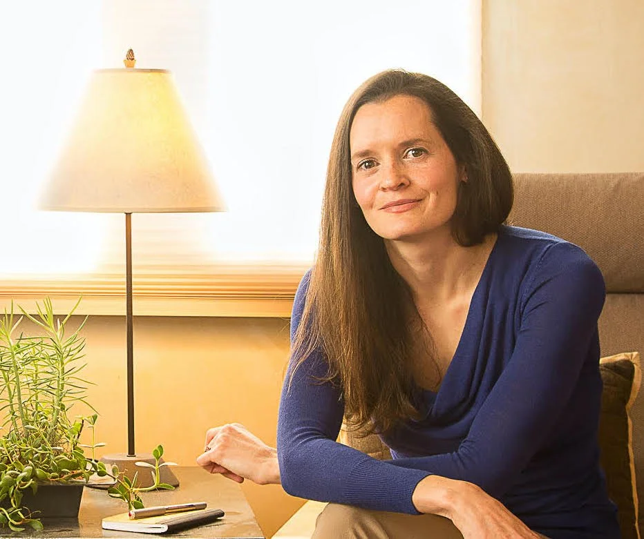 A woman with long brown hair and a blue shirt sitting on a beige couch in a well-lit room with a lamp and green houseplant on a side table.