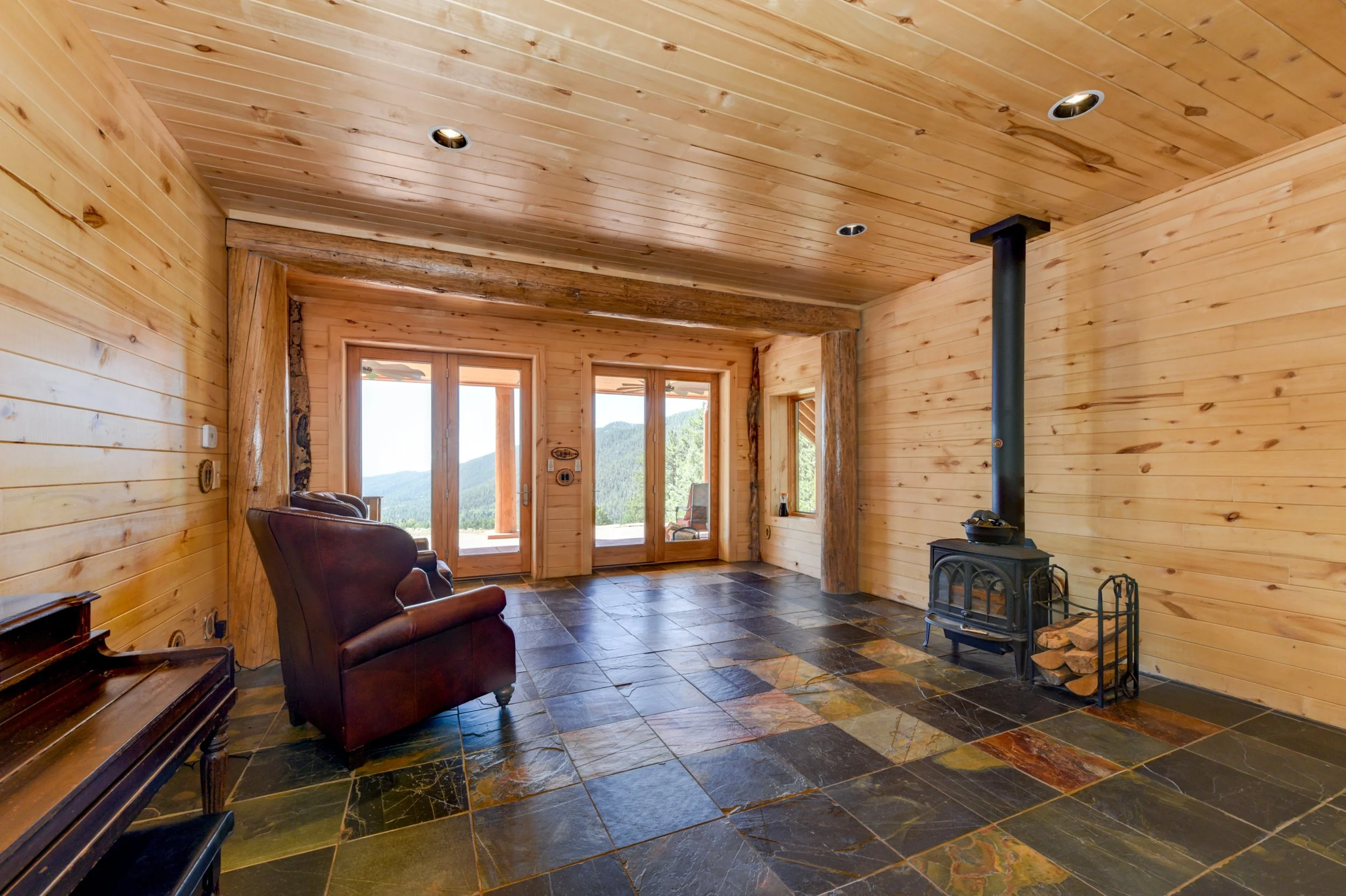Living room with wooden walls and ceiling, slate tile floor, leather armchair, wood-burning stove, and large windows with mountain views.