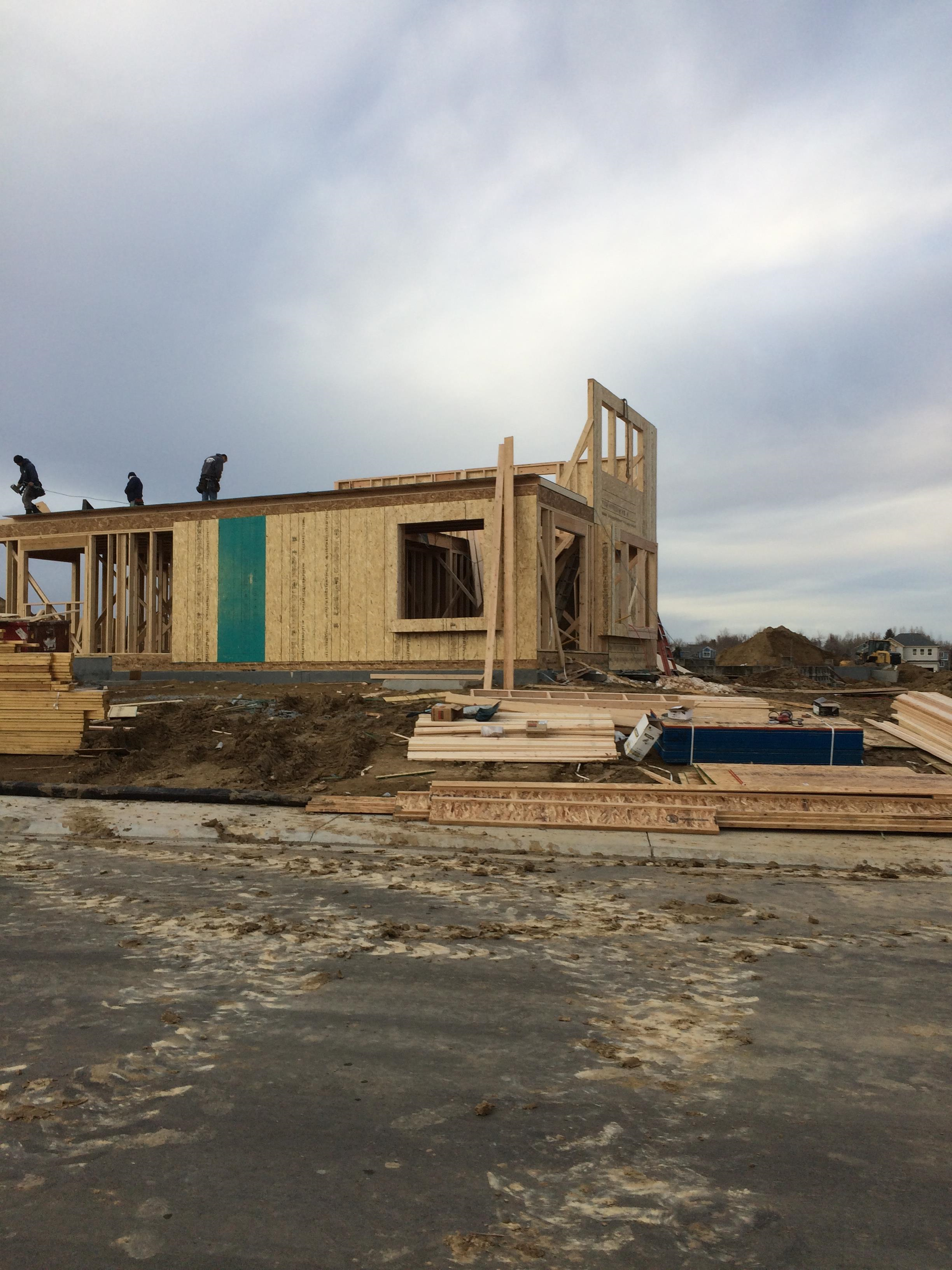 Construction site with partially built house and workers on the roof, wooden framing, building materials, and dirt ground.