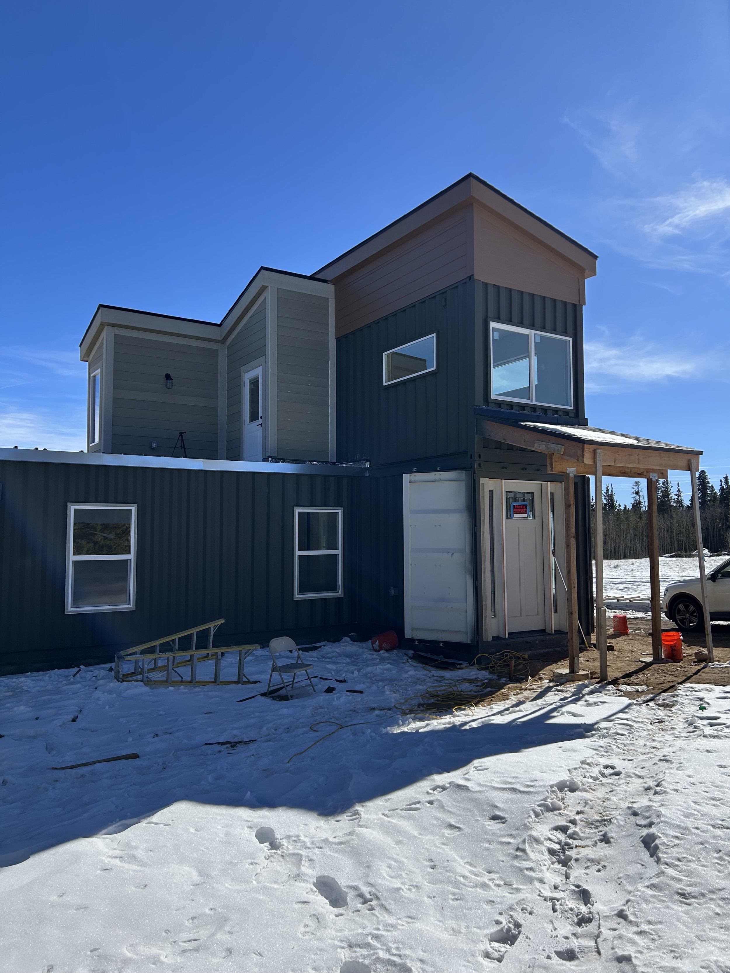 A multi-story shipping container house under construction with snow on the ground, exposed construction materials, and a clear blue sky.