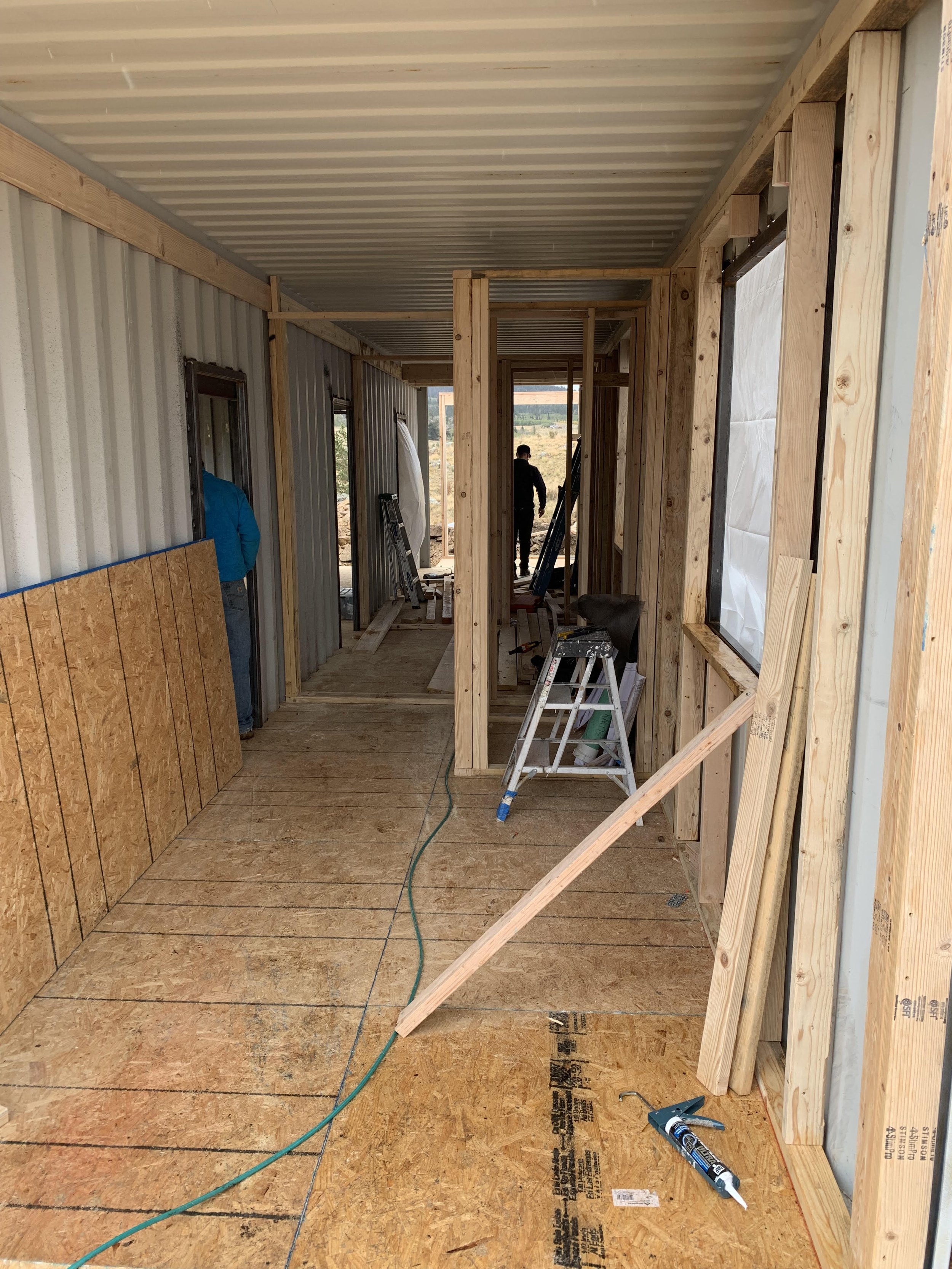 Interior of a building under construction, showing wood framing, plywood floors, and construction tools, with two workers in the background.