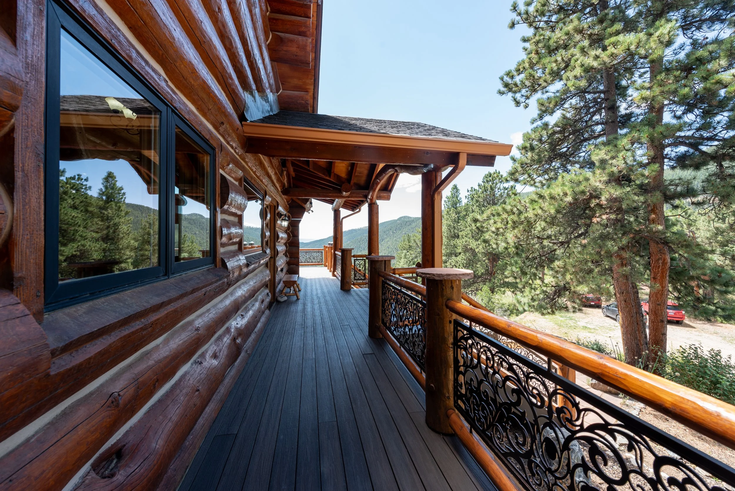 Wooden cabin balcony with dark railing, surrounded by green pine trees, mountain landscape in the distance, bright blue sky.