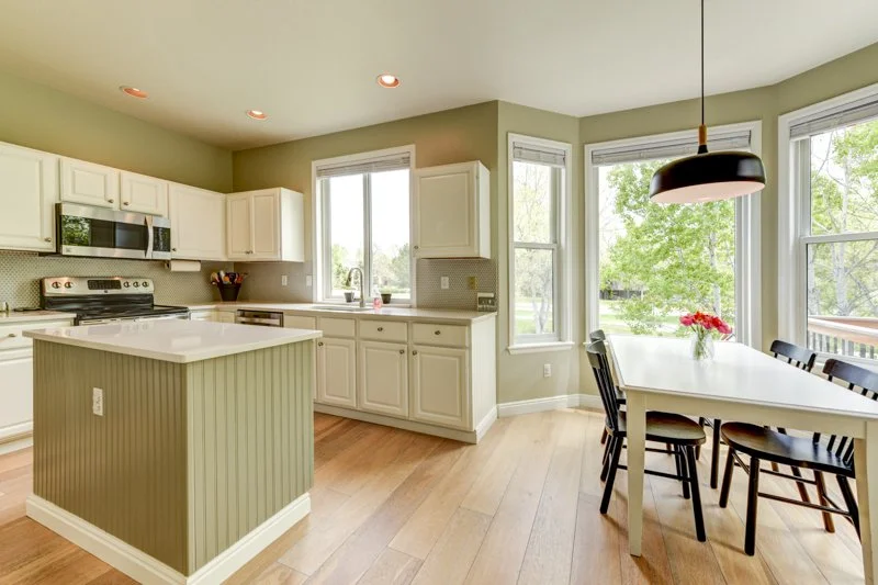 Bright kitchen with white cabinets, a central island, and a dining area with a white table, black chairs, and a hanging light fixture; large windows showing green trees outside.